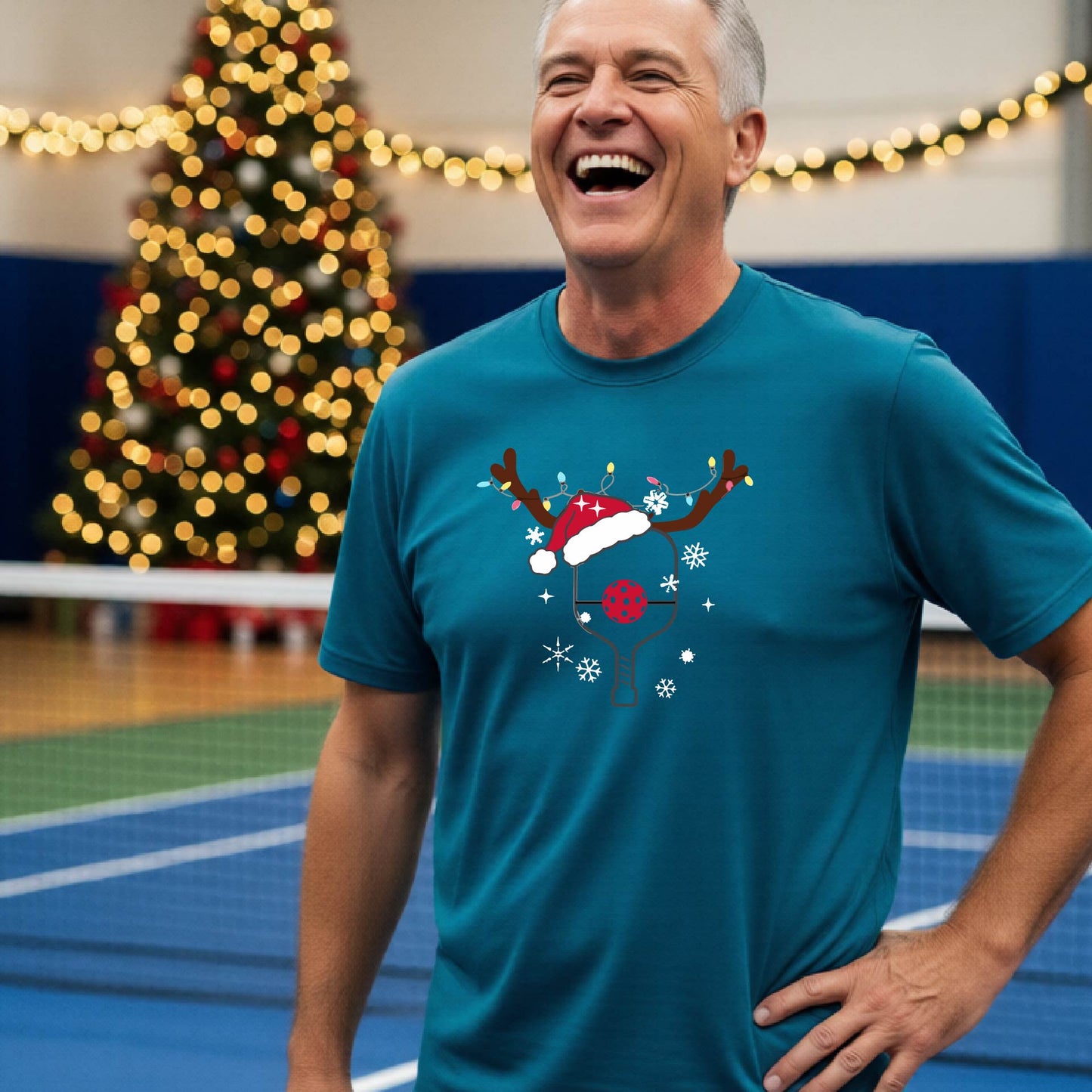 A man wearing a teal graphic t-shirt with a Christmas-themed pickleball design featuring a pickleball paddle wearing a Santa hat and reindeer antlers, and snowflakes, set against a blurred background of a pickleball court and holiday lights.