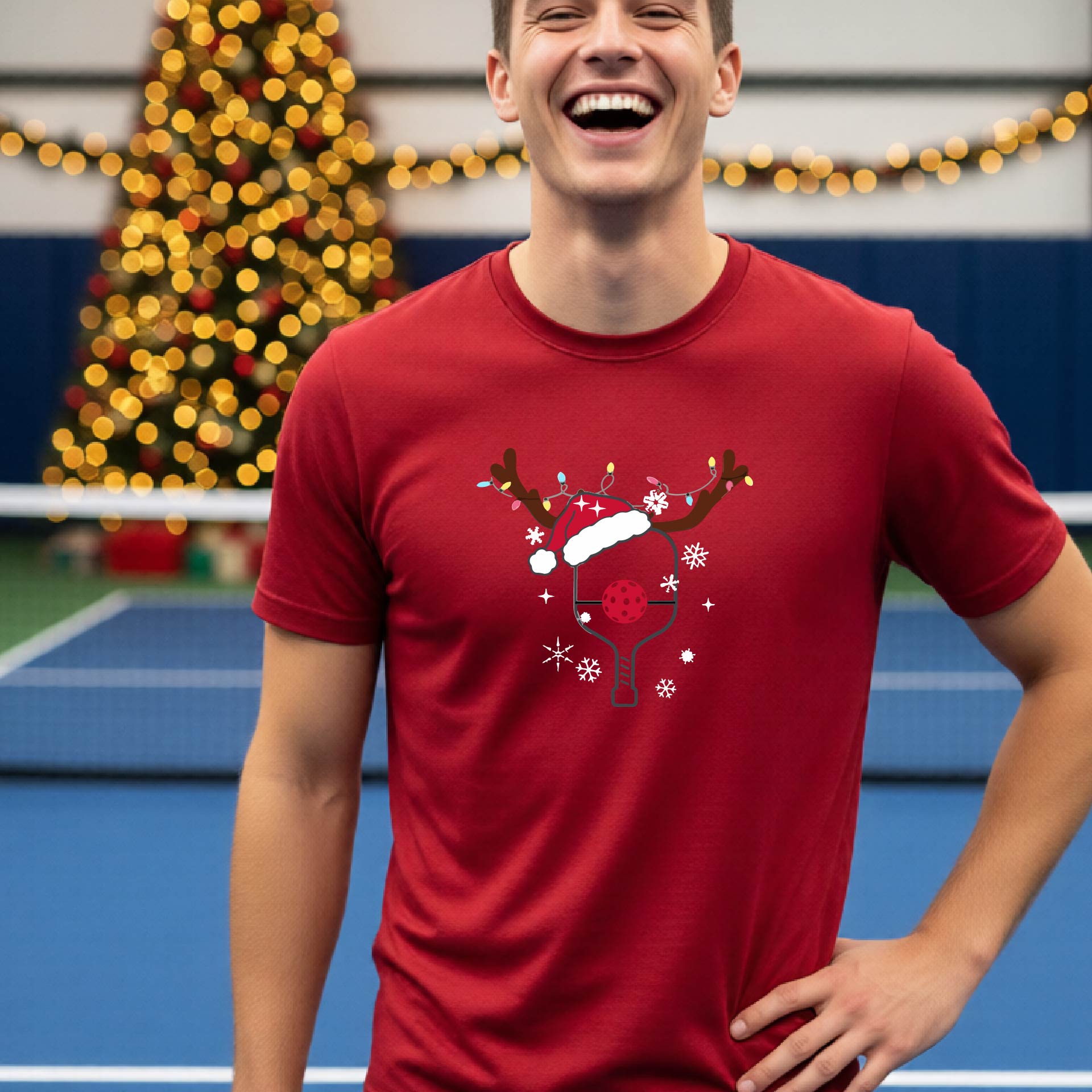 A man wearing a black graphic t-shirt with a Christmas-themed pickleball design featuring a pickleball paddle wearing a Santa hat and reindeer antlers, and snowflakes, set against a blurred background of a pickleball court and holiday lights.