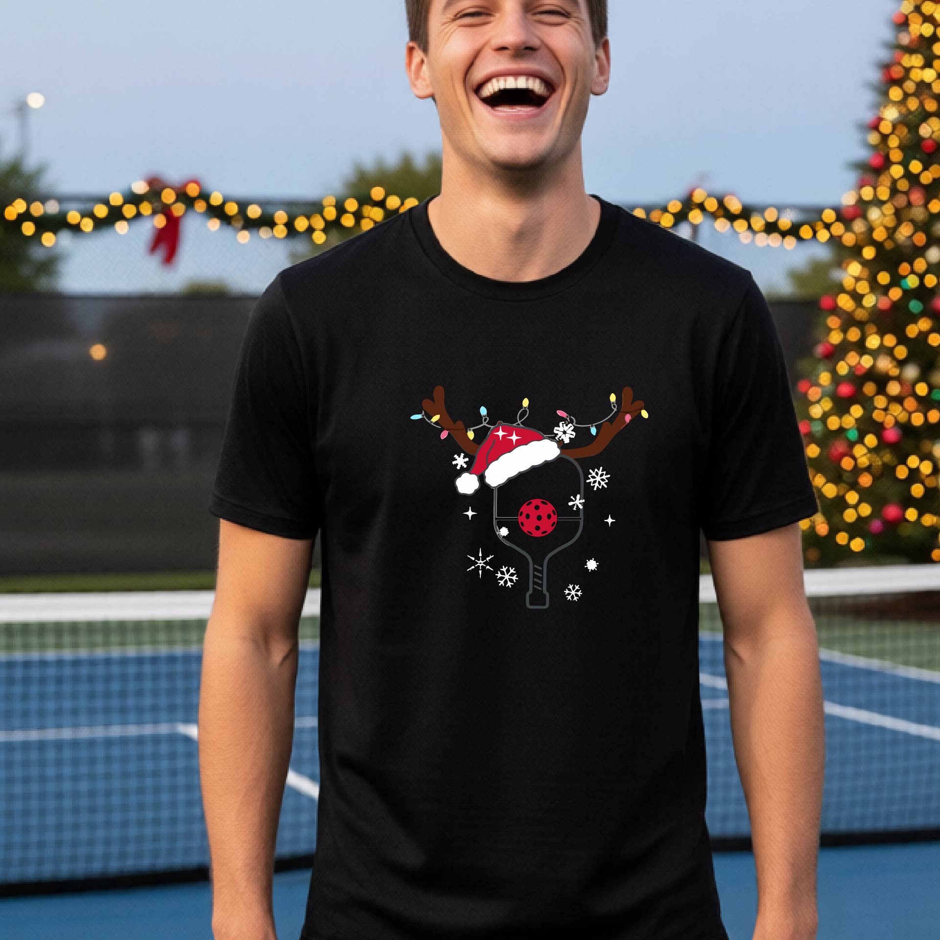 A man wearing a black graphic t-shirt with a Christmas-themed pickleball design featuring a pickleball paddle wearing a Santa hat, and snowflakes, set against a blurred background of a pickleball court and holiday lights.
