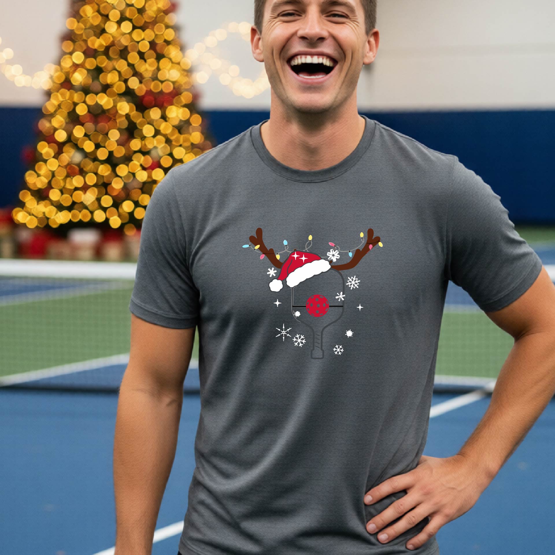 A man wearing a gray graphic t-shirt with a Christmas-themed pickleball design featuring a pickleball paddle wearing a Santa hat, and snowflakes, set against a blurred background of a pickleball court and holiday lights.