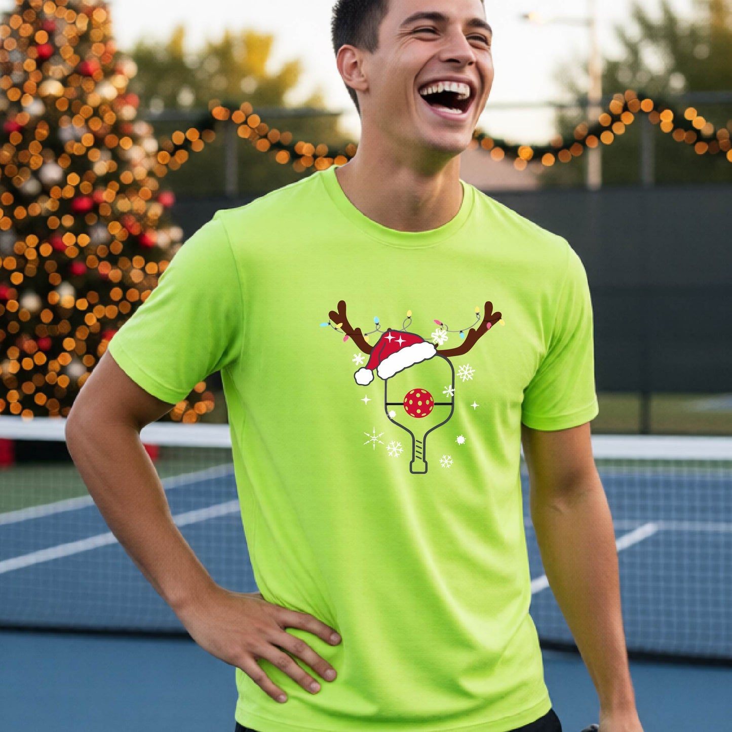 A man wearing a lime green graphic t-shirt with a Christmas-themed pickleball design featuring a pickleball paddle wearing a Santa hat, and snowflakes, set against a blurred background of a pickleball court and holiday lights.