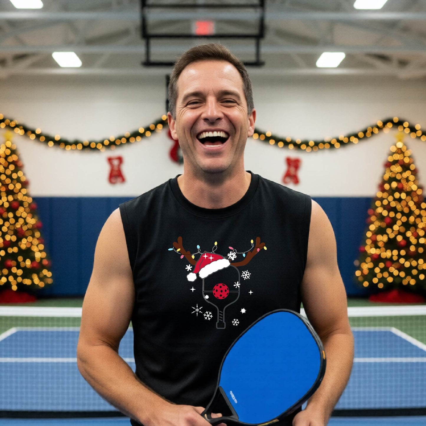 A smiling man wearing a black sleeveless shirt with a festive Christmas-themed pickleball graphic of a pickleball paddle, pickleball, and reindeer antlers, standing on a pickleball court with string lights and a decorated Christmas tree in the background.