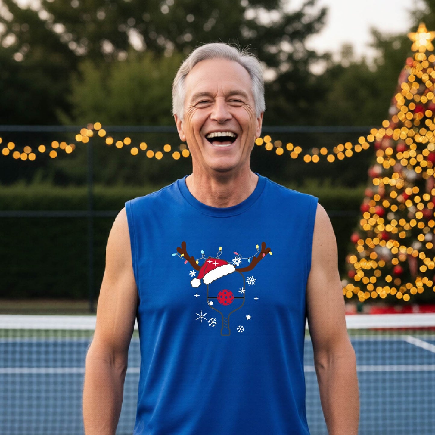 A smiling man wearing a royal blue sleeveless shirt with a festive Christmas-themed pickleball graphic of a pickleball paddle, pickleball, and reindeer antlers, standing on a pickleball court with string lights and a decorated Christmas tree in the background.