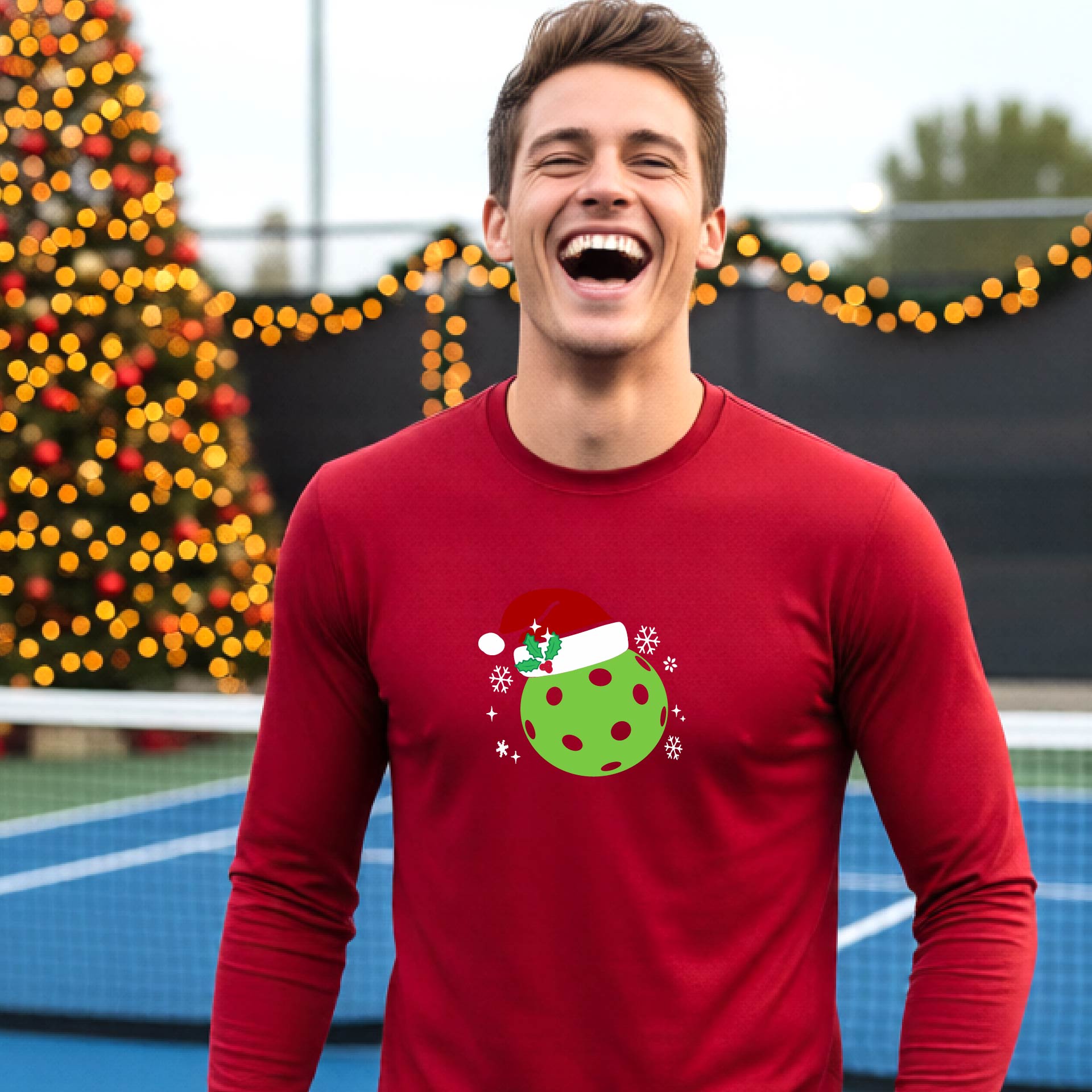 A man wearing a red long-sleeve shirt with a Christmas-themed graphic of a green pickleball wearing a red Santa hat, set against a background of a decorated Christmas tree and a pickleball court.