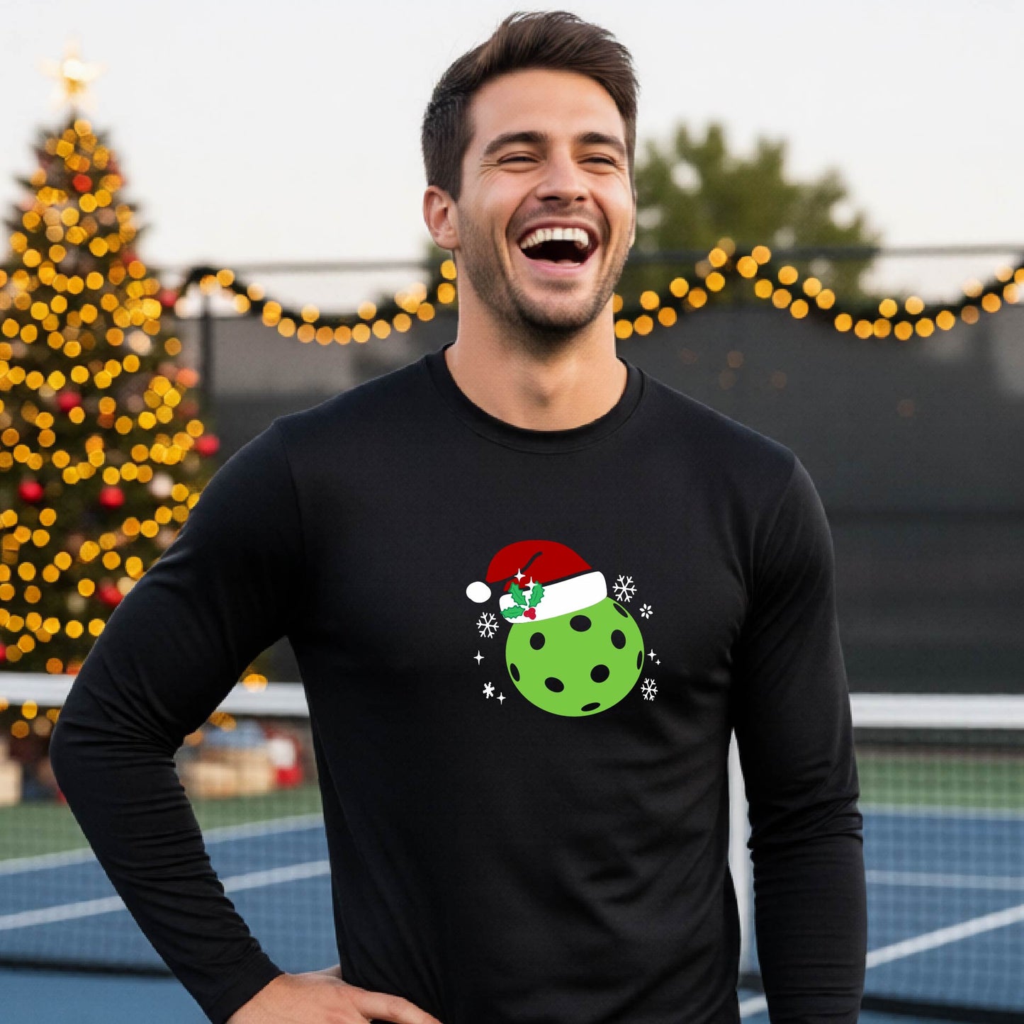 A man wearing a black long-sleeve shirt with a Christmas-themed graphic of a green pickleball wearing a red Santa hat, set against a background of a decorated Christmas tree and a pickleball court.