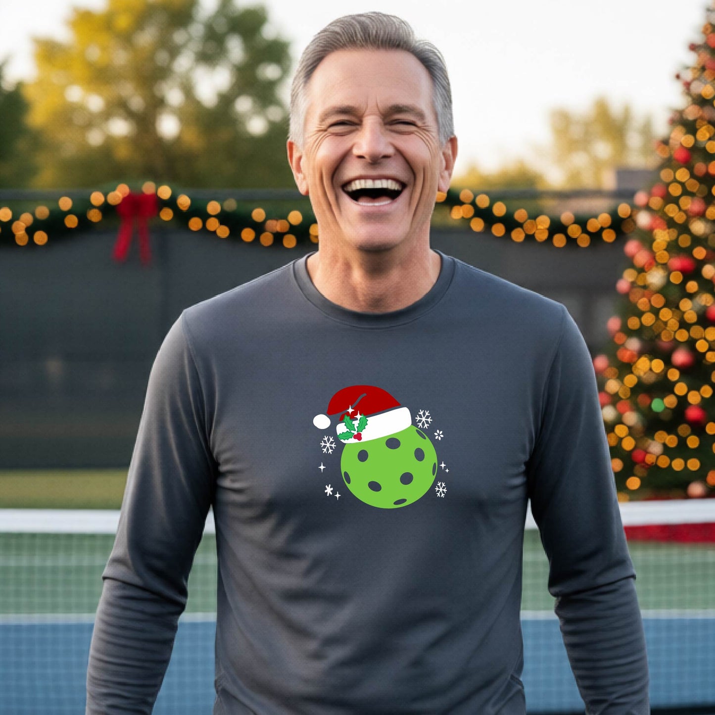 A man wearing a gray long-sleeve shirt with a Christmas-themed graphic of a green pickleball wearing a red Santa hat, set against a background of a decorated Christmas tree and a pickleball court.