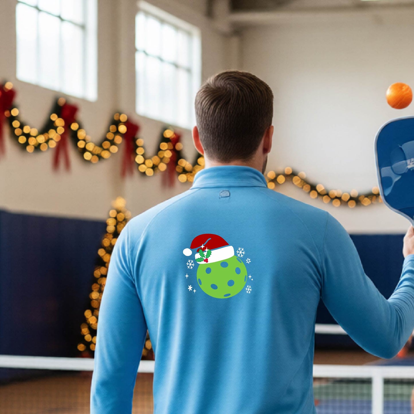 A man viewed from behind wearing a light blue long-sleeve quarter-zip pullover with a Christmas-themed graphic on the back. The graphic features a green pickkleball with black polka dots wearing a red Santa hat, surrounded by small white snowflakes.