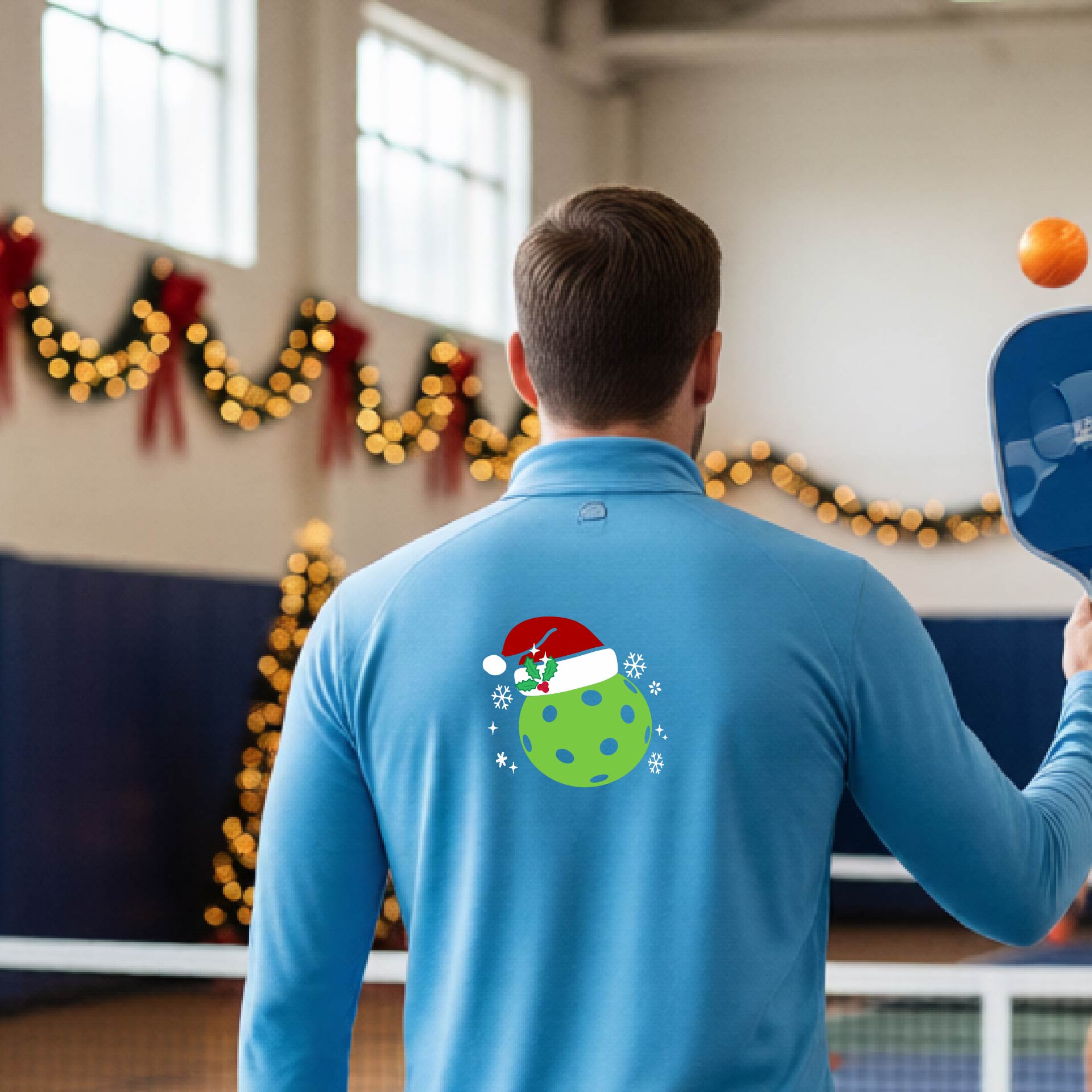 A man viewed from behind wearing a light blue long-sleeve quarter-zip pullover with a Christmas-themed graphic on the back. The graphic features a green pickkleball with black polka dots wearing a red Santa hat, surrounded by small white snowflakes.