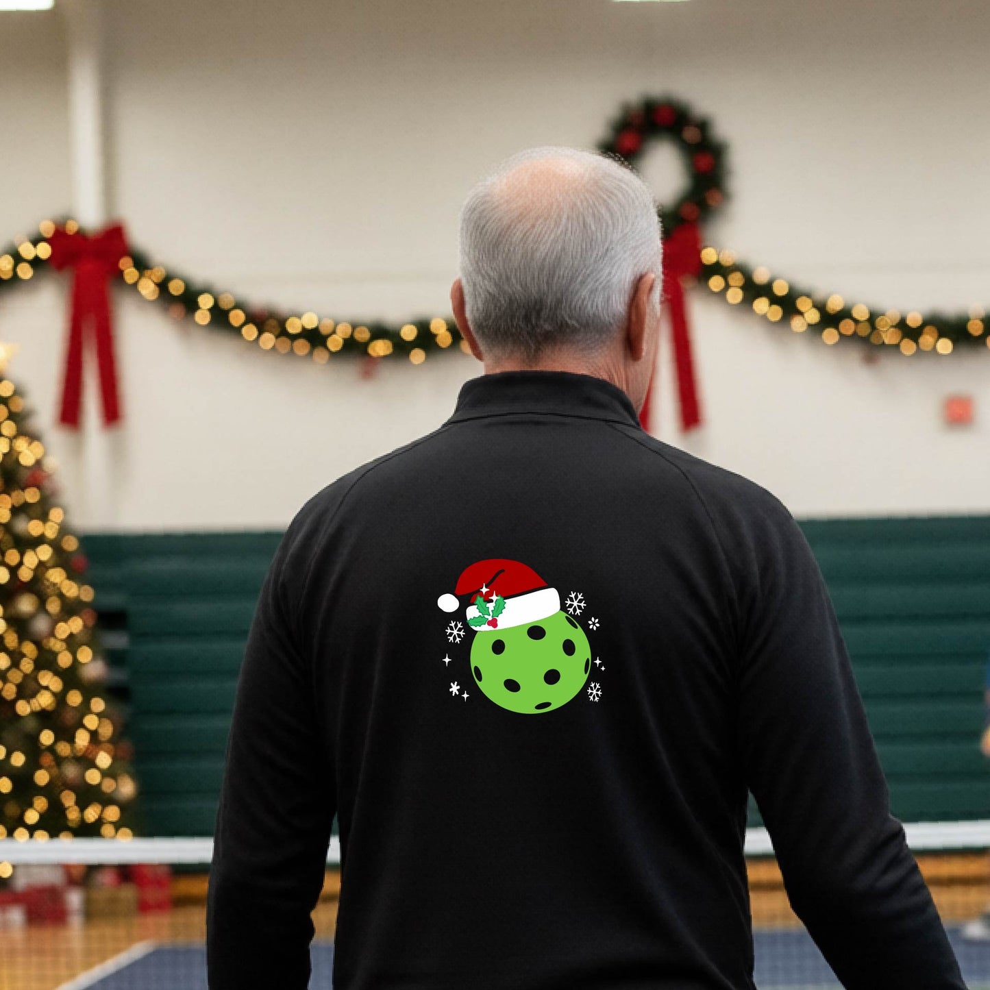 A man viewed from behind wearing a black long-sleeve quarter-zip pullover with a Christmas-themed graphic on the back. The graphic features a green pickkleball with black polka dots wearing a red Santa hat, surrounded by small white snowflakes.