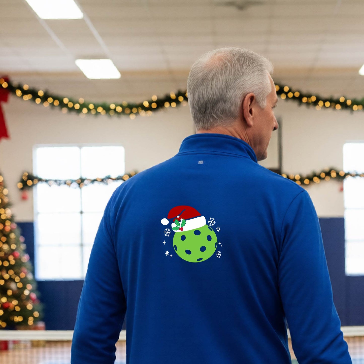 A man viewed from behind wearing a royal blue long-sleeve quarter-zip pullover with a Christmas-themed graphic on the back. The graphic features a green pickkleball with black polka dots wearing a red Santa hat, surrounded by small white snowflakes.