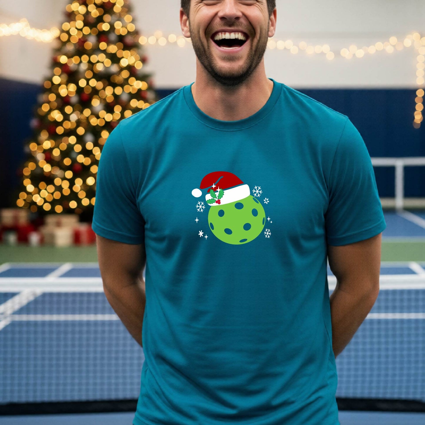 A man wearing a teal blue short-sleeve t-shirt with a Christmas pickleball wearing a Santahat graphic, standing on an indoor tennis court with a blurred Christmas tree in the background.