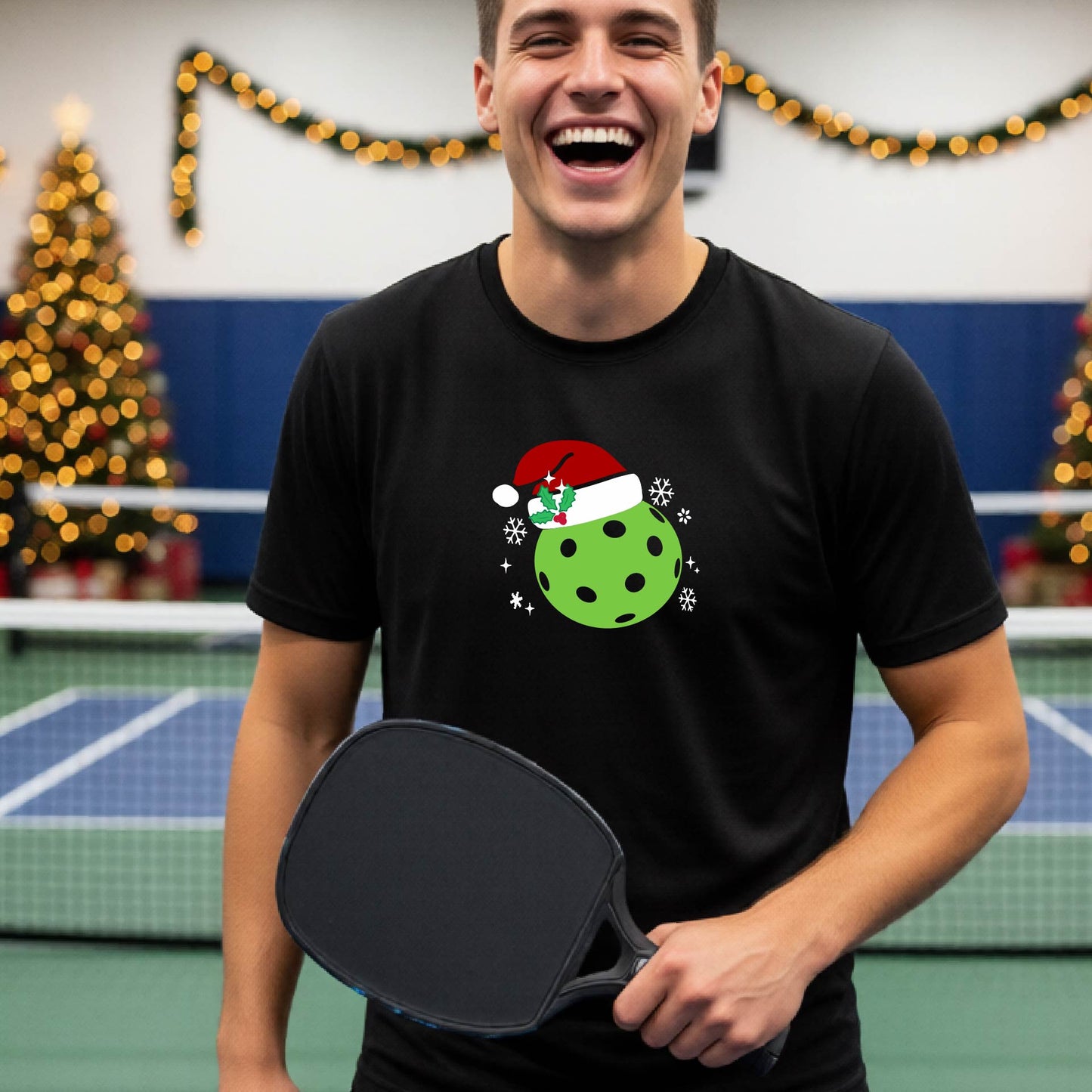 A man wearing a black short-sleeve t-shirt with a Christmas pickleball wearing a Santahat graphic, standing on an indoor tennis court with a blurred Christmas tree in the background.