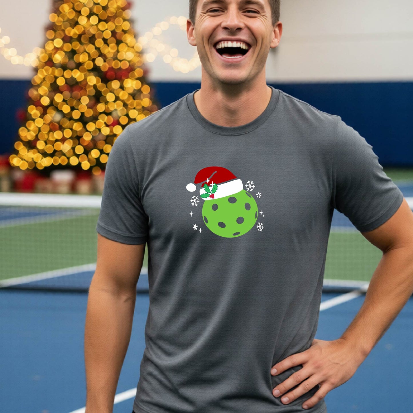 A man wearing a gray short-sleeve t-shirt with a Christmas pickleball wearing a Santahat graphic, standing on an indoor tennis court with a blurred Christmas tree in the background.