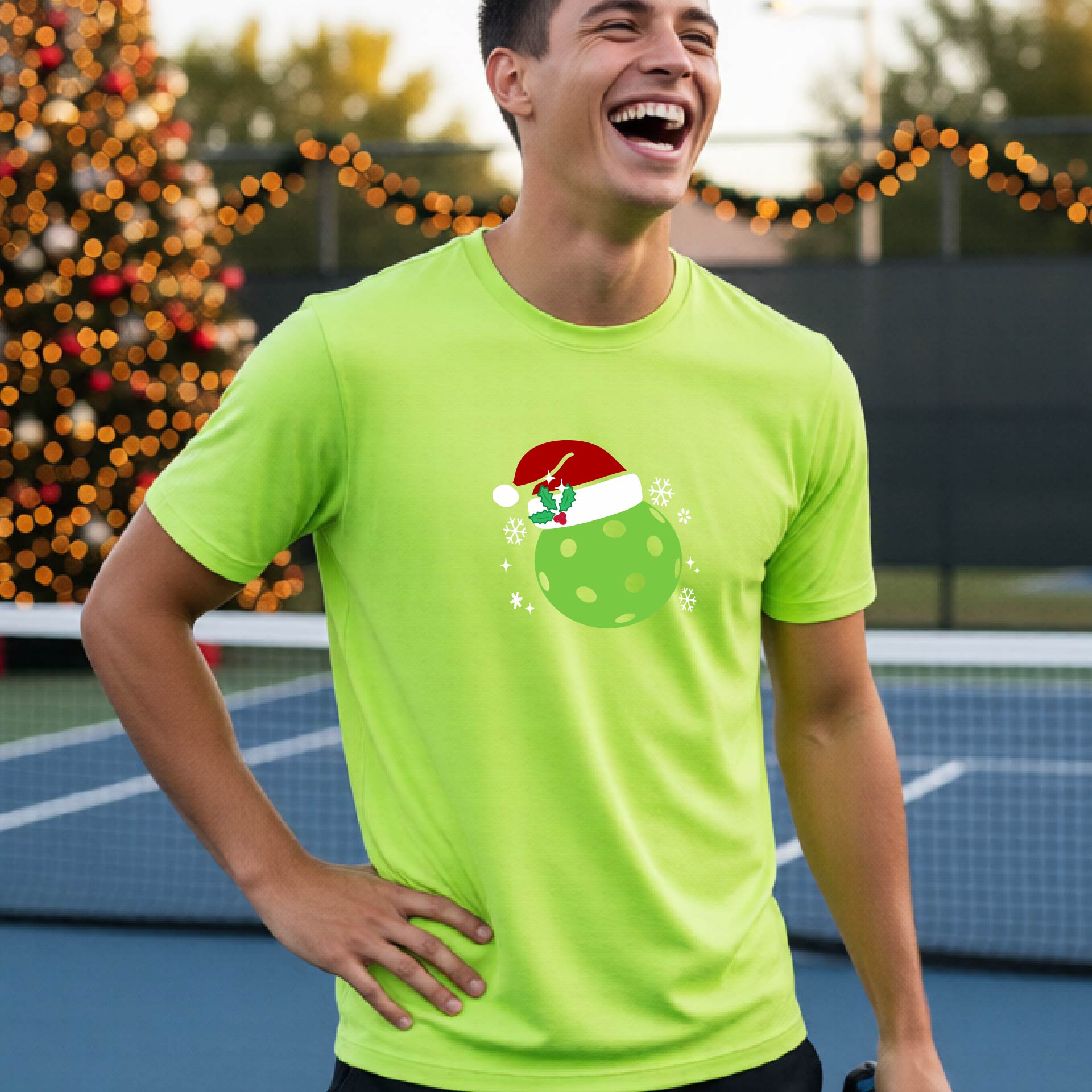 A man wearing a lime green short-sleeve t-shirt with a Christmas pickleball wearing a Santahat graphic, standing on an indoor tennis court with a blurred Christmas tree in the background.