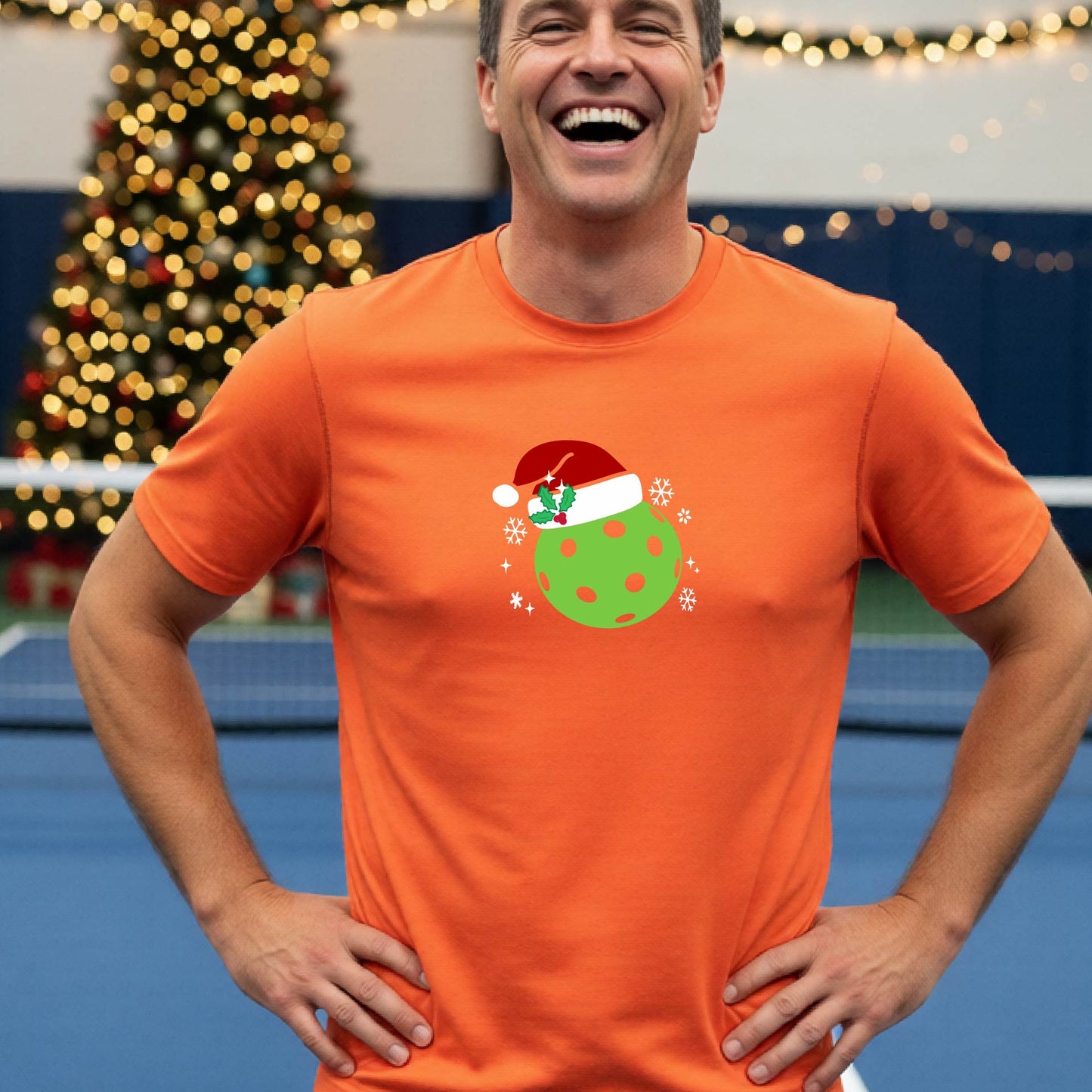 A man wearing a bright orange short-sleeve t-shirt with a Christmas pickleball wearing a Santahat graphic, standing on an indoor tennis court with a blurred Christmas tree in the background.