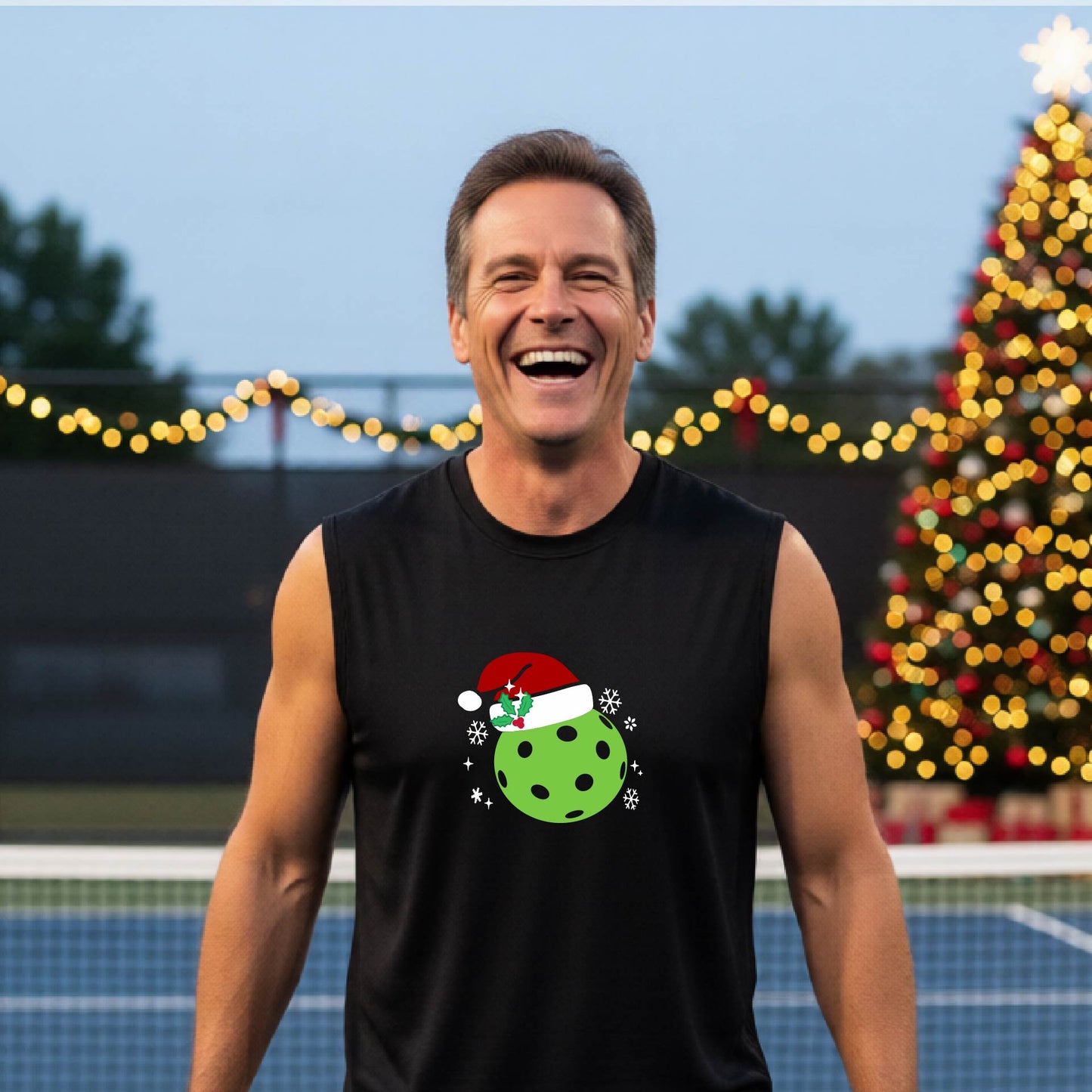 A smiling man wearing a black sleeveless shirt with a green pickleball wearing a Santa hat Christmas-themed graphic stands on a pickleball court, with a lit Christmas tree and warm string lights in the background.