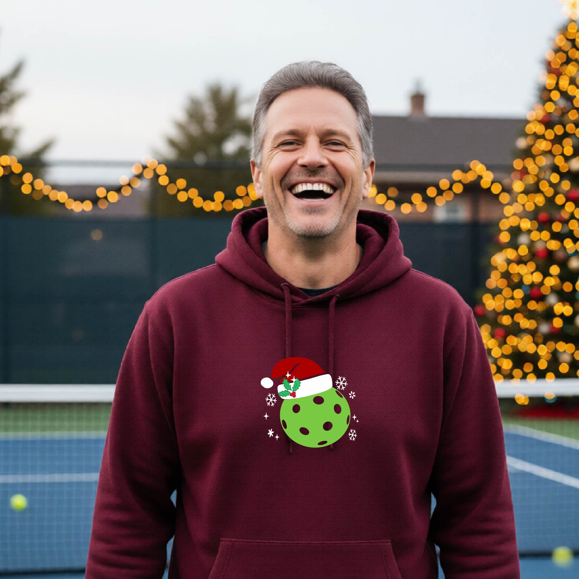 An man wearing a maroon hoodie with a green Christmas-themed pickleball wearing a Santa hat graphic on the chest, on a pickleball court decorated with holiday lights.