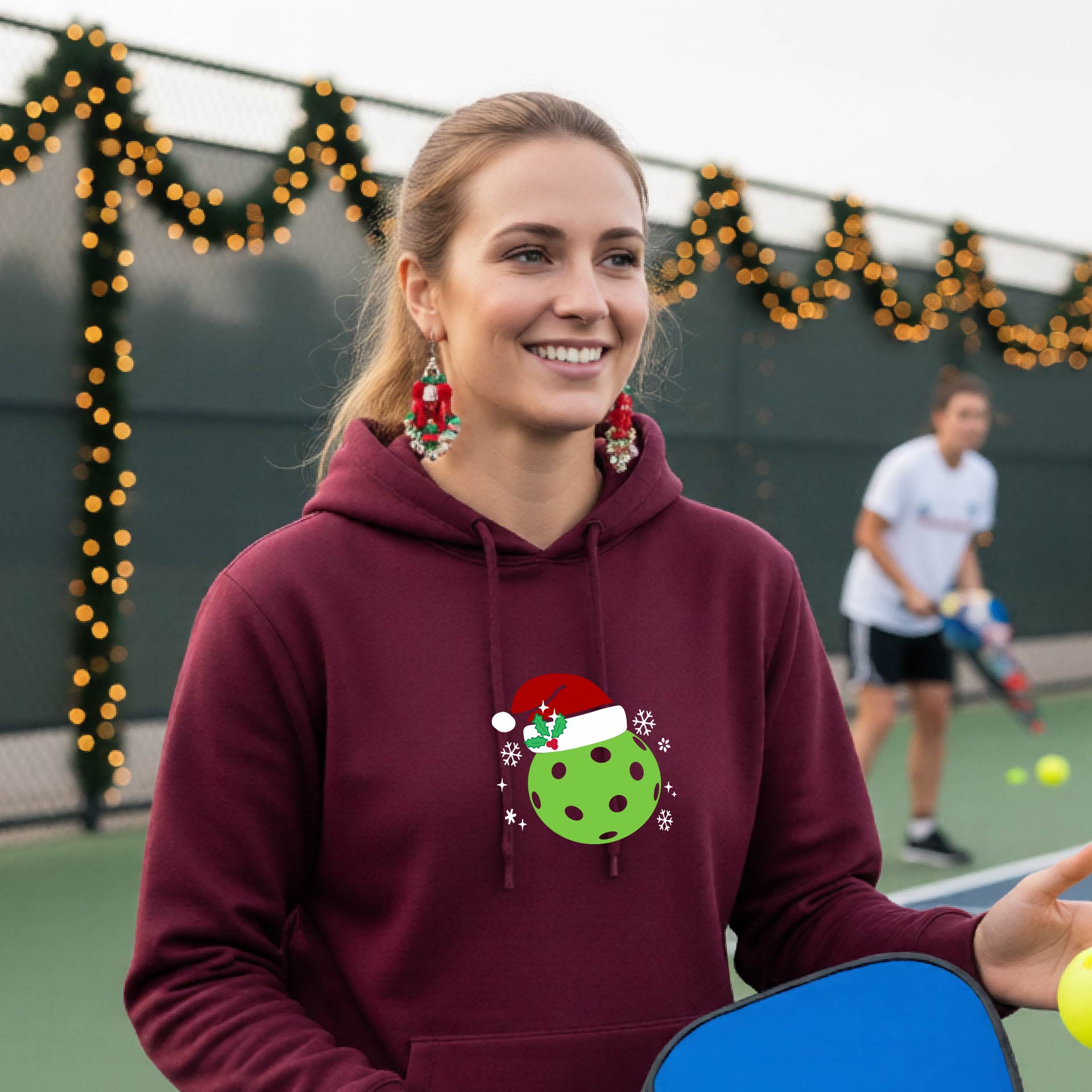 An woman wearing a maroon hoodie with a green Christmas-themed pickleball wearing a Santa hat graphic on the chest, on a pickleball court decorated with holiday lights.