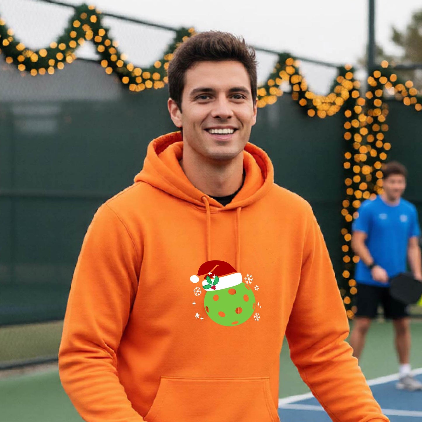 An man wearing a bright orange hoodie with a green Christmas-themed pickleball wearing a Santa hat graphic on the chest, on a pickleball court decorated with holiday lights.