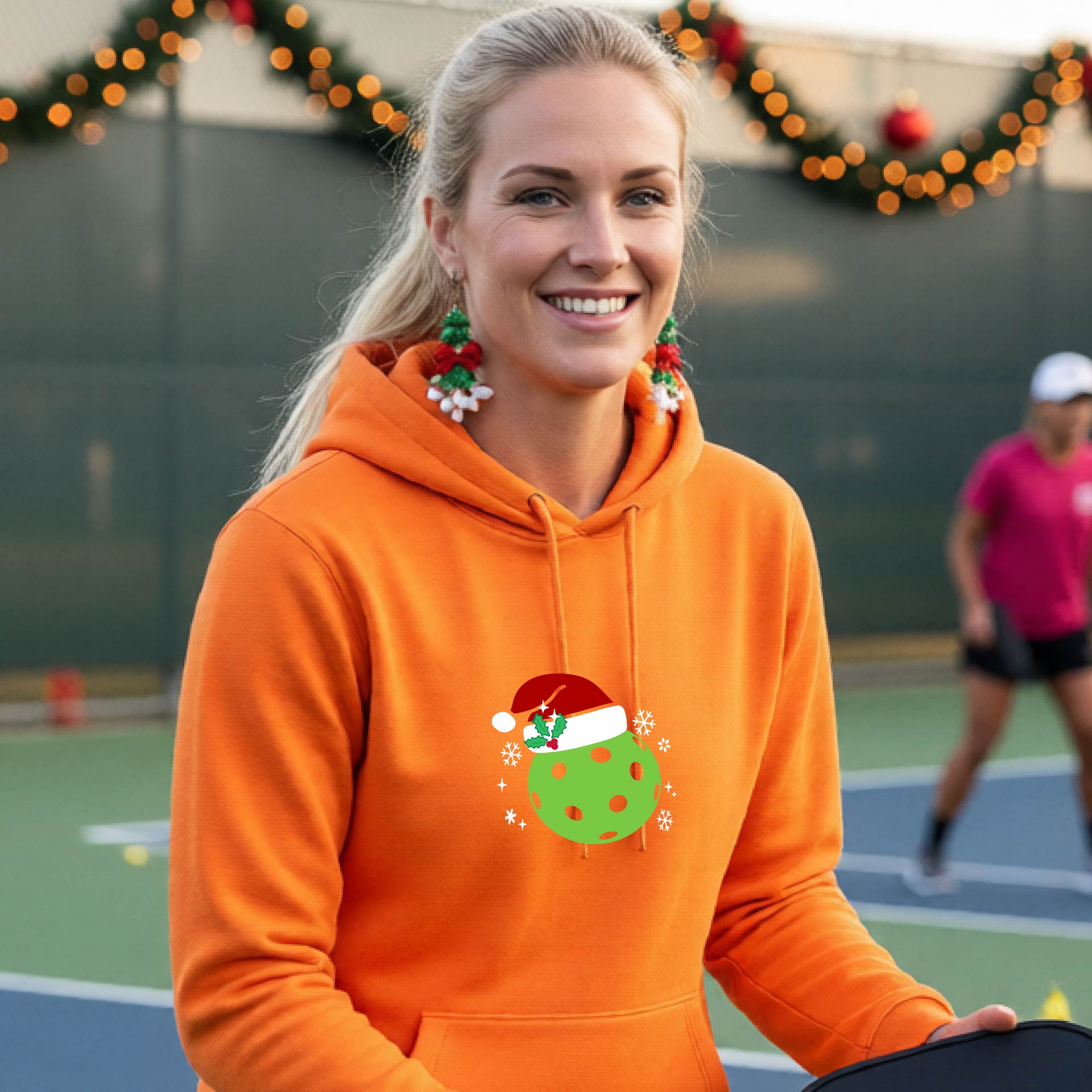 An woman wearing a bright orange hoodie with a green Christmas-themed pickleball wearing a Santa hat graphic on the chest, on a pickleball court decorated with holiday lights.