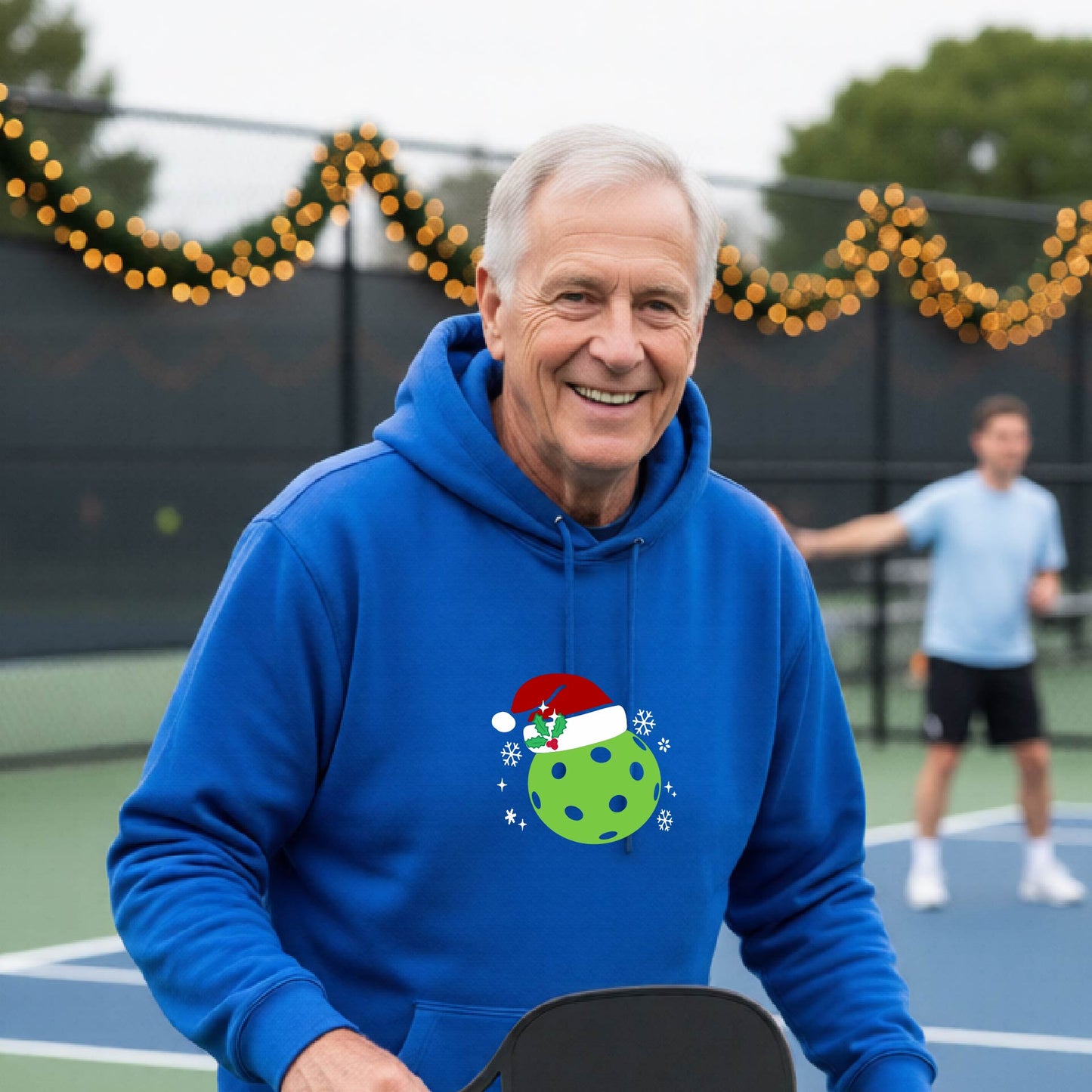 An man wearing a royal blue hoodie with a green Christmas-themed pickleball wearing a Santa hat graphic on the chest, on a pickleball court decorated with holiday lights.