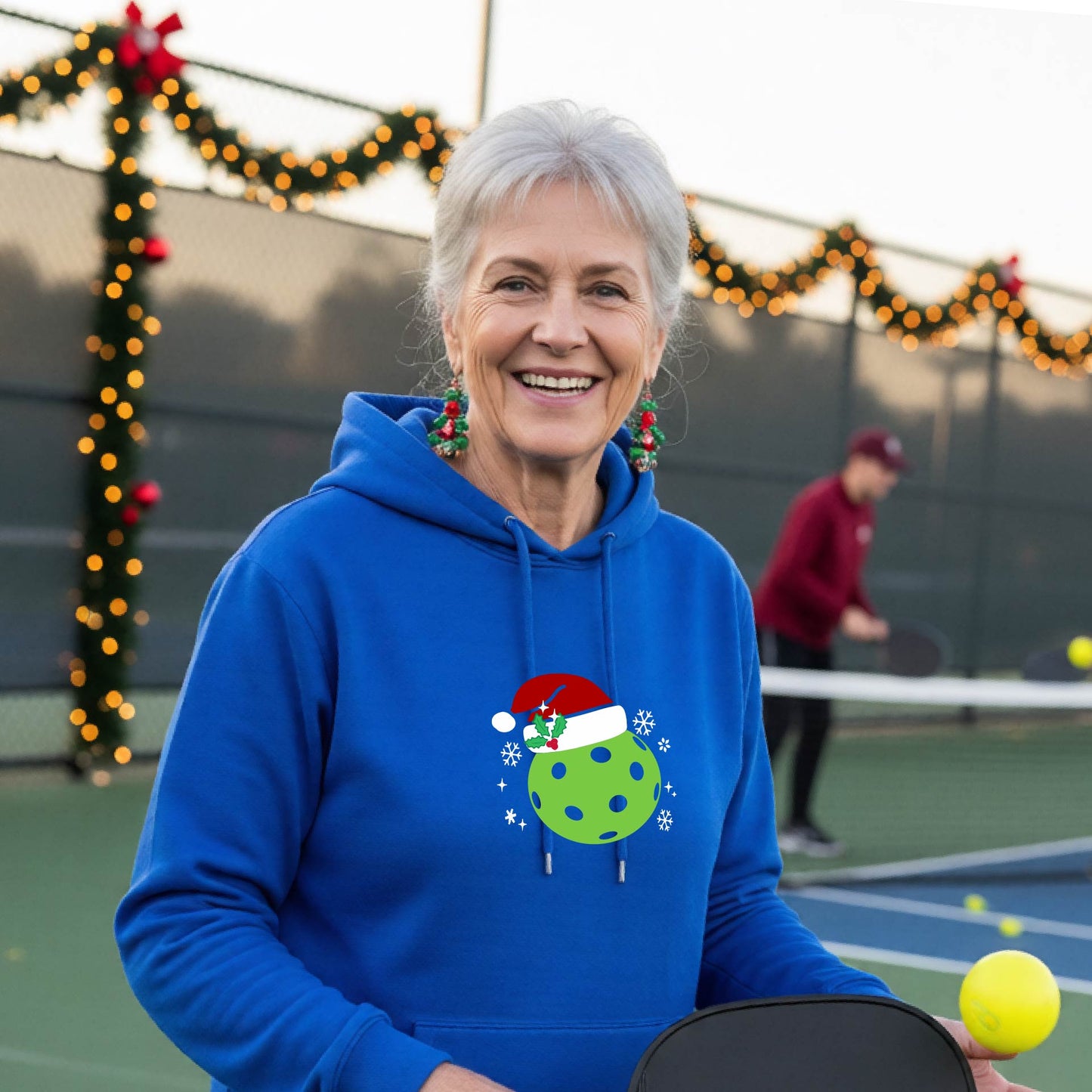 Anwo man wearing a royal blue hoodie with a green Christmas-themed pickleball wearing a Santa hat graphic on the chest, on a pickleball court decorated with holiday lights.