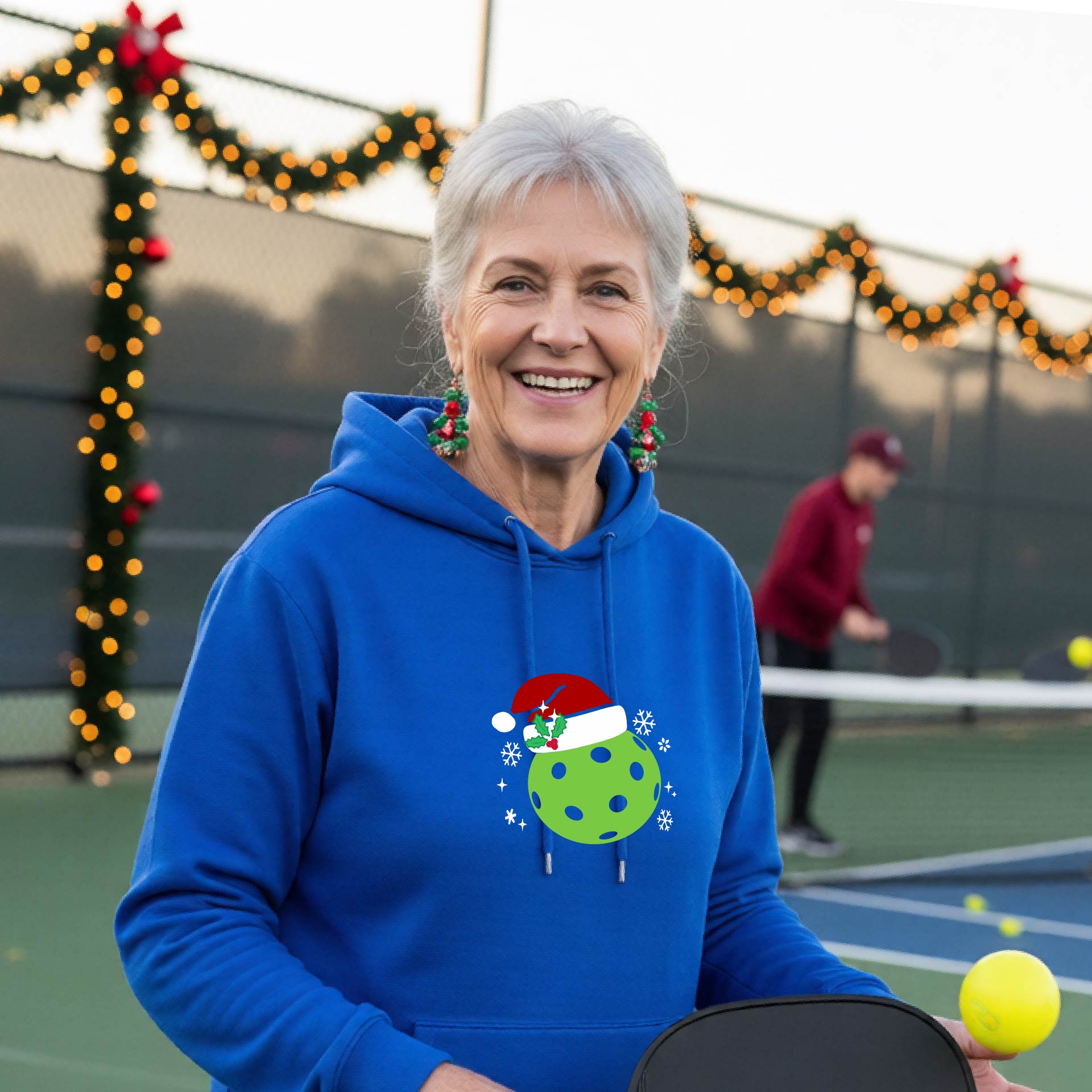 Anwo man wearing a royal blue hoodie with a green Christmas-themed pickleball wearing a Santa hat graphic on the chest, on a pickleball court decorated with holiday lights.