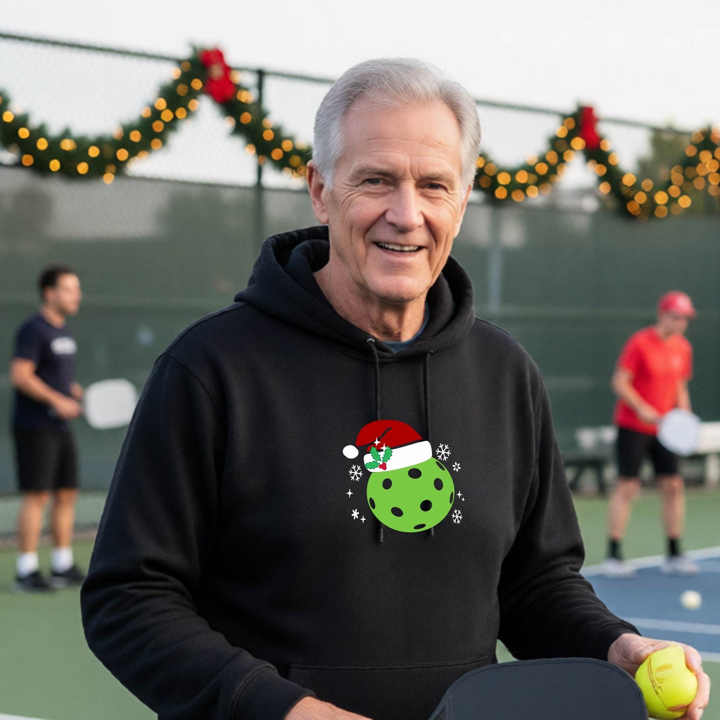 An man wearing a black hoodie with a green Christmas-themed pickleball wearing a Santa hat graphic on the chest, on a pickleball court decorated with holiday lights.