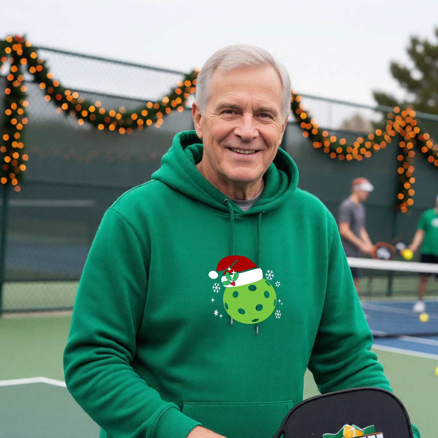 An man wearing a green hoodie with a green Christmas-themed pickleball wearing a Santa hat graphic on the chest, on a pickleball court decorated with holiday lights.