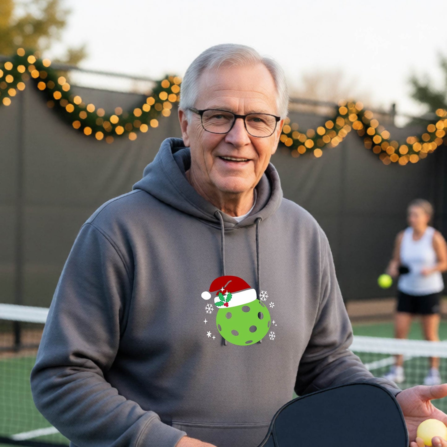 An man wearing a gray hoodie with a green Christmas-themed pickleball wearing a Santa hat graphic on the chest, on a pickleball court decorated with holiday lights.