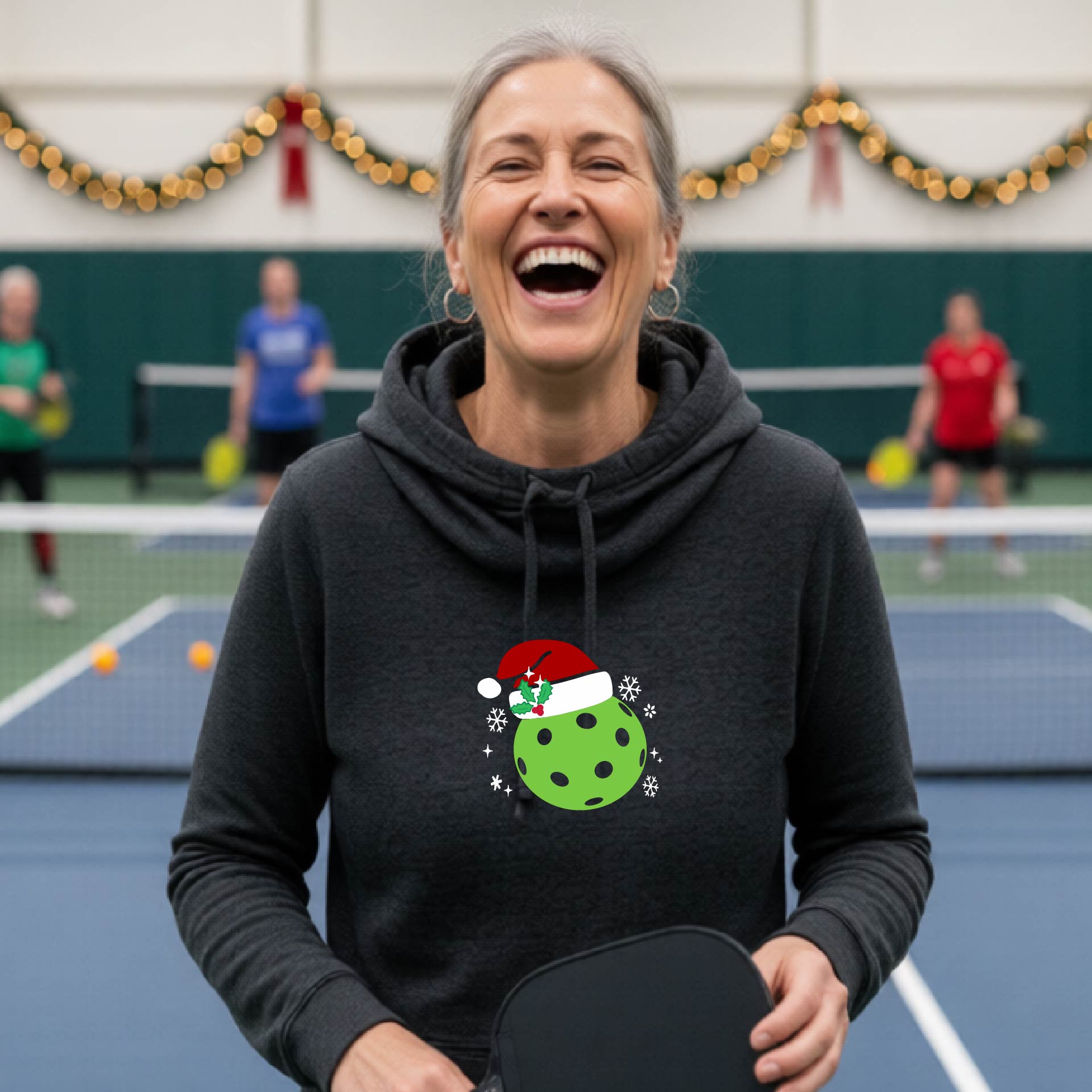 A heather black cowl-neck hooded sweatshirt featuring a pickleball graphic of a green pickleball wearing a santa hat and Christmas themed background. 