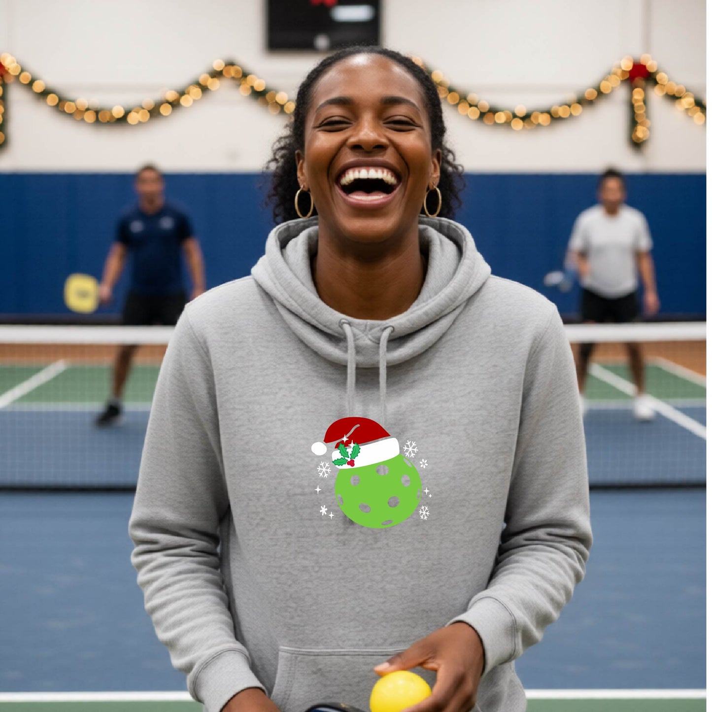 A grey cowl-neck hooded sweatshirt featuring a pickleball graphic of a green pickleball wearing a santa hat and Christmas themed background. 