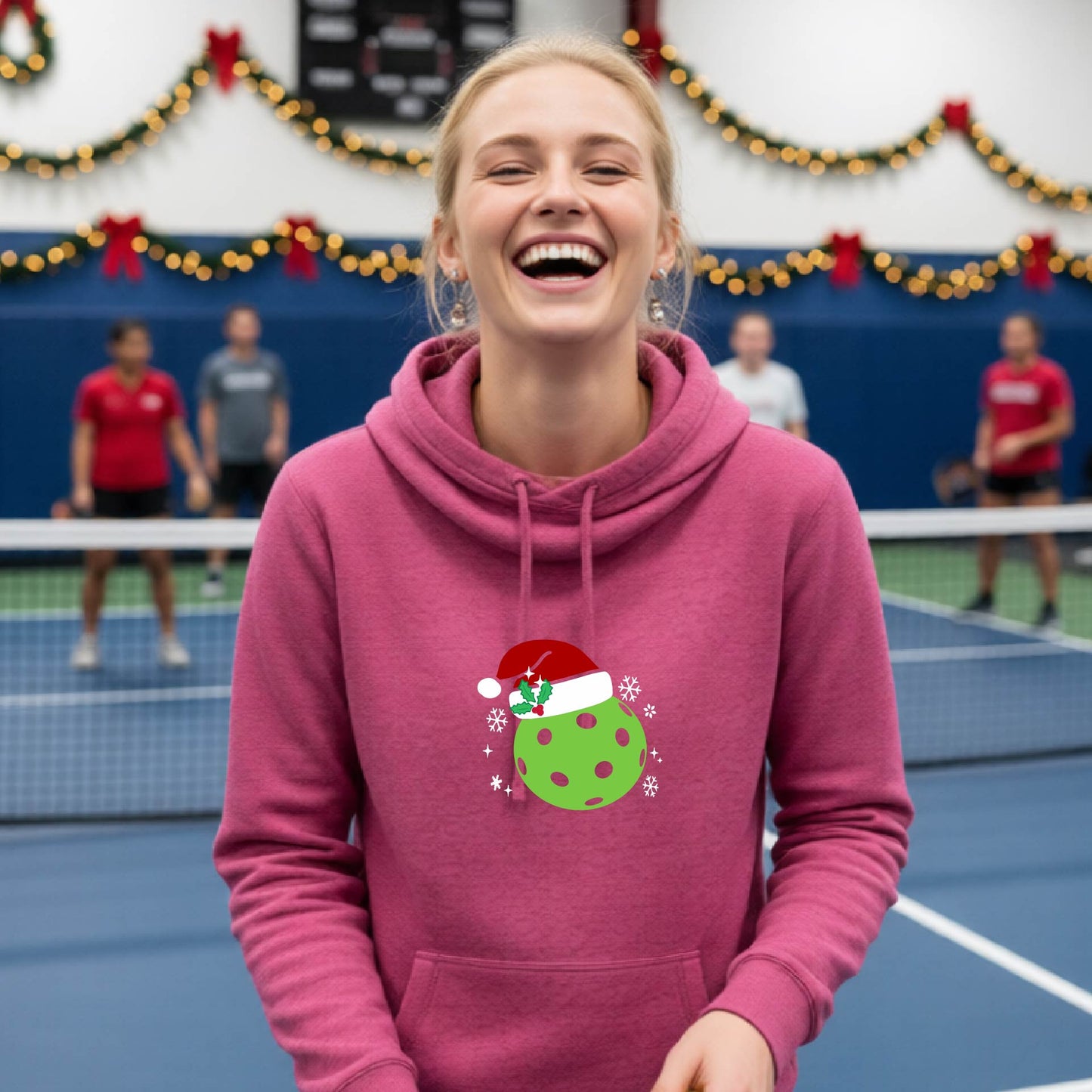 A pink cowl-neck hooded sweatshirt featuring a pickleball graphic of a green pickleball wearing a santa hat and Christmas themed background. 