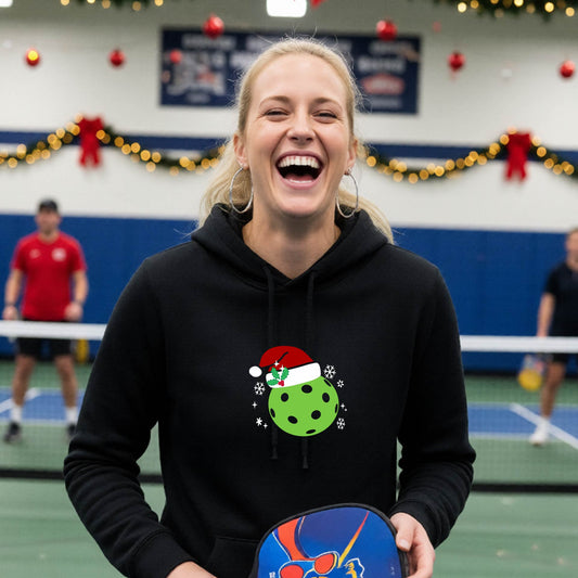 A black hooded sweatshirt featuring a pickleball graphic with a green pickleball wearing a santa hat and a Christmas themed background. 