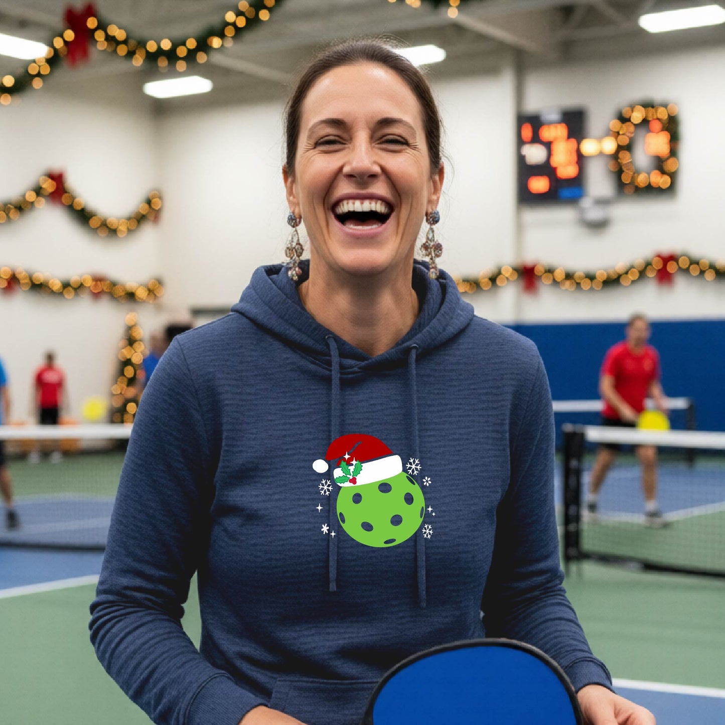 A navy blue hooded sweatshirt featuring a pickleball graphic with a green pickleball wearing a santa hat and a Christmas themed background. 