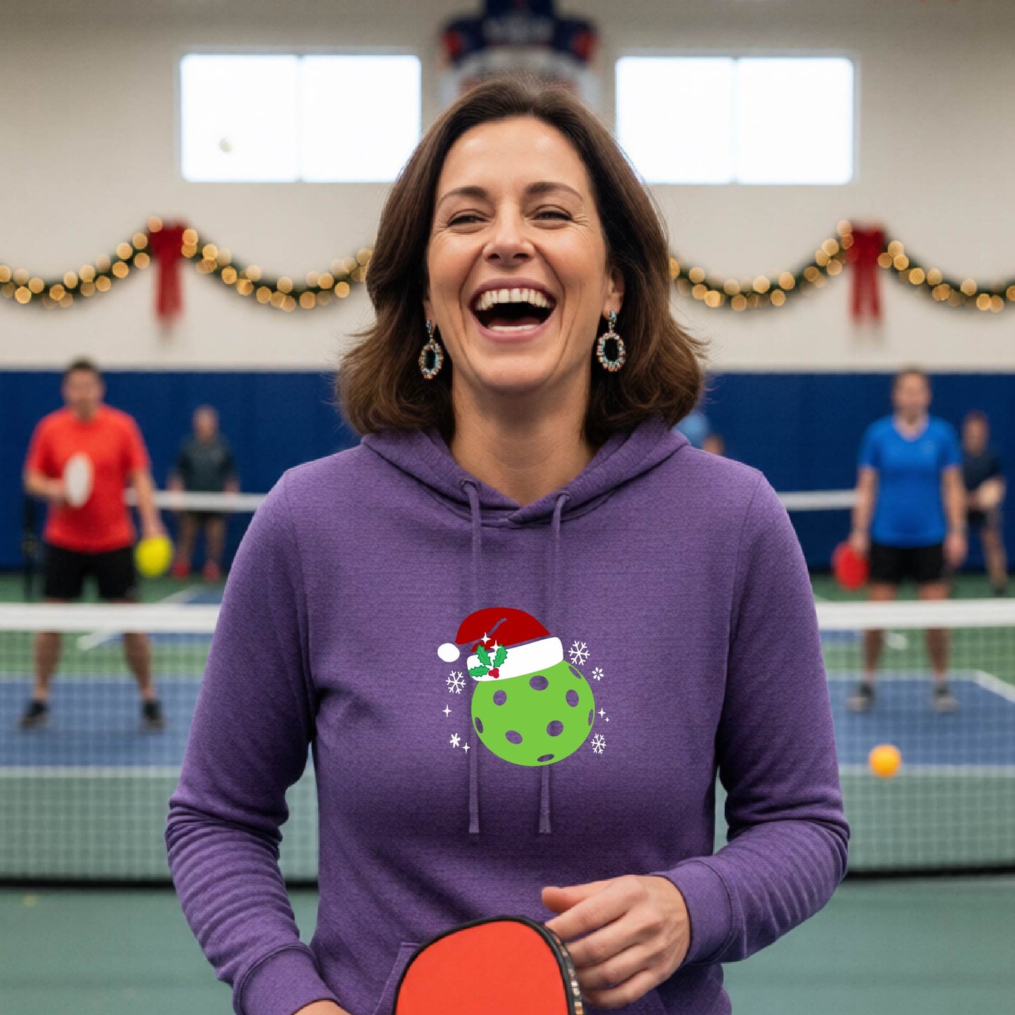 A purple hooded sweatshirt featuring a pickleball graphic with a green pickleball wearing a santa hat and a Christmas themed background. 