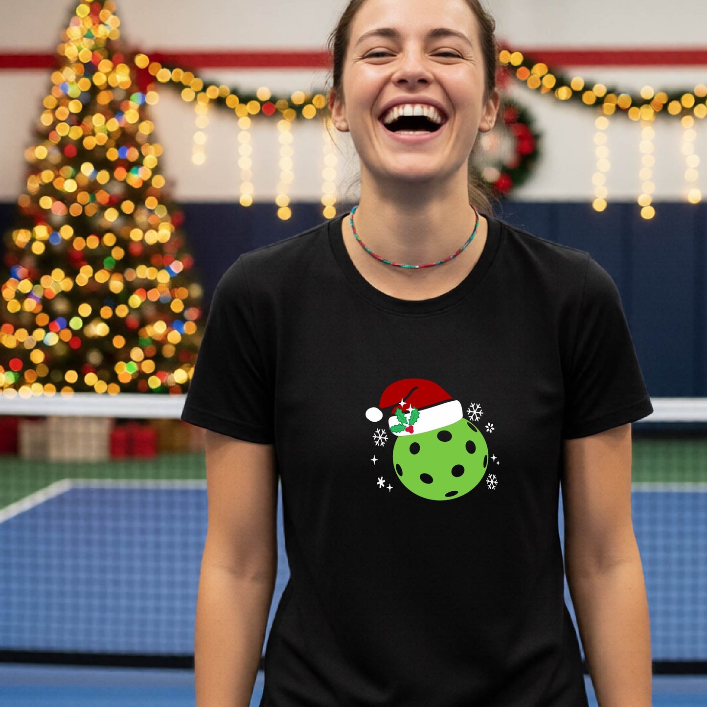 A woman wearing a black graphic t-shirt with a green Christmas-themed pickleball design, standing in front of a blurred Christmas tree and holiday decorations.