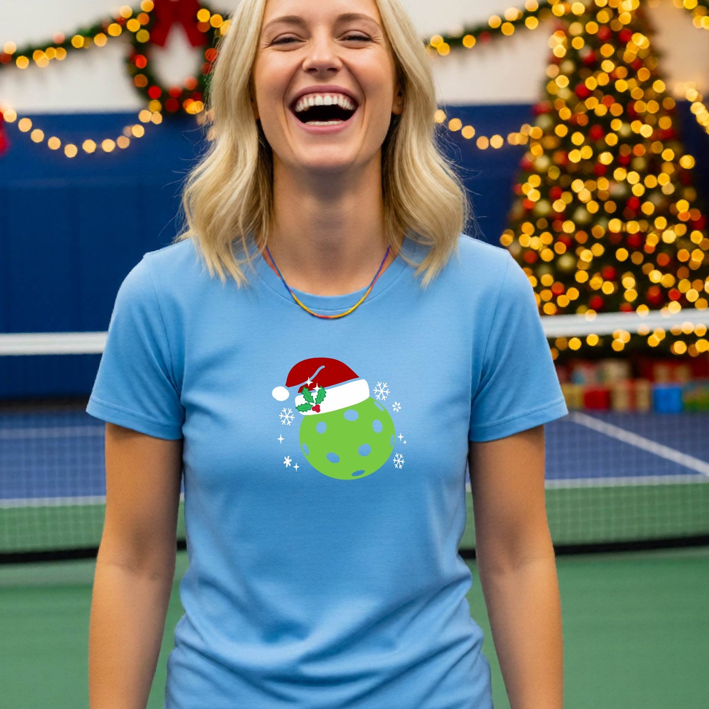 A woman wearing a light blue graphic t-shirt with a green Christmas-themed pickleball design, standing in front of a blurred Christmas tree and holiday decorations.