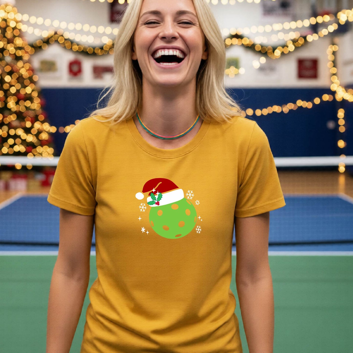A woman wearing a mustard yellow gold graphic t-shirt with a green Christmas-themed pickleball design, standing in front of a blurred Christmas tree and holiday decorations.