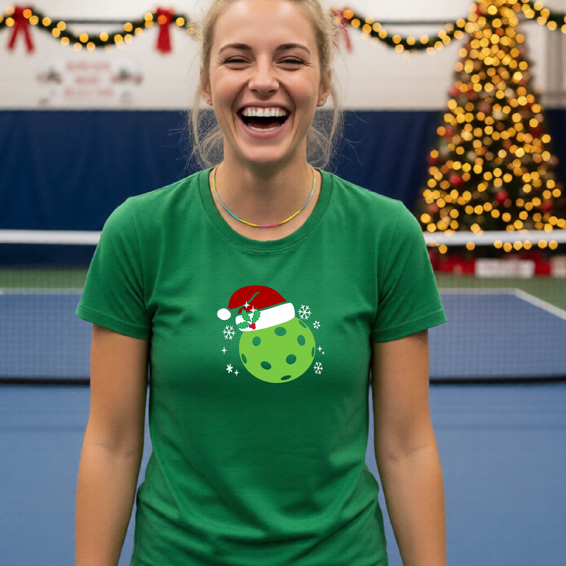 A woman wearing a green graphic t-shirt with a green Christmas-themed pickleball design, standing in front of a blurred Christmas tree and holiday decorations.