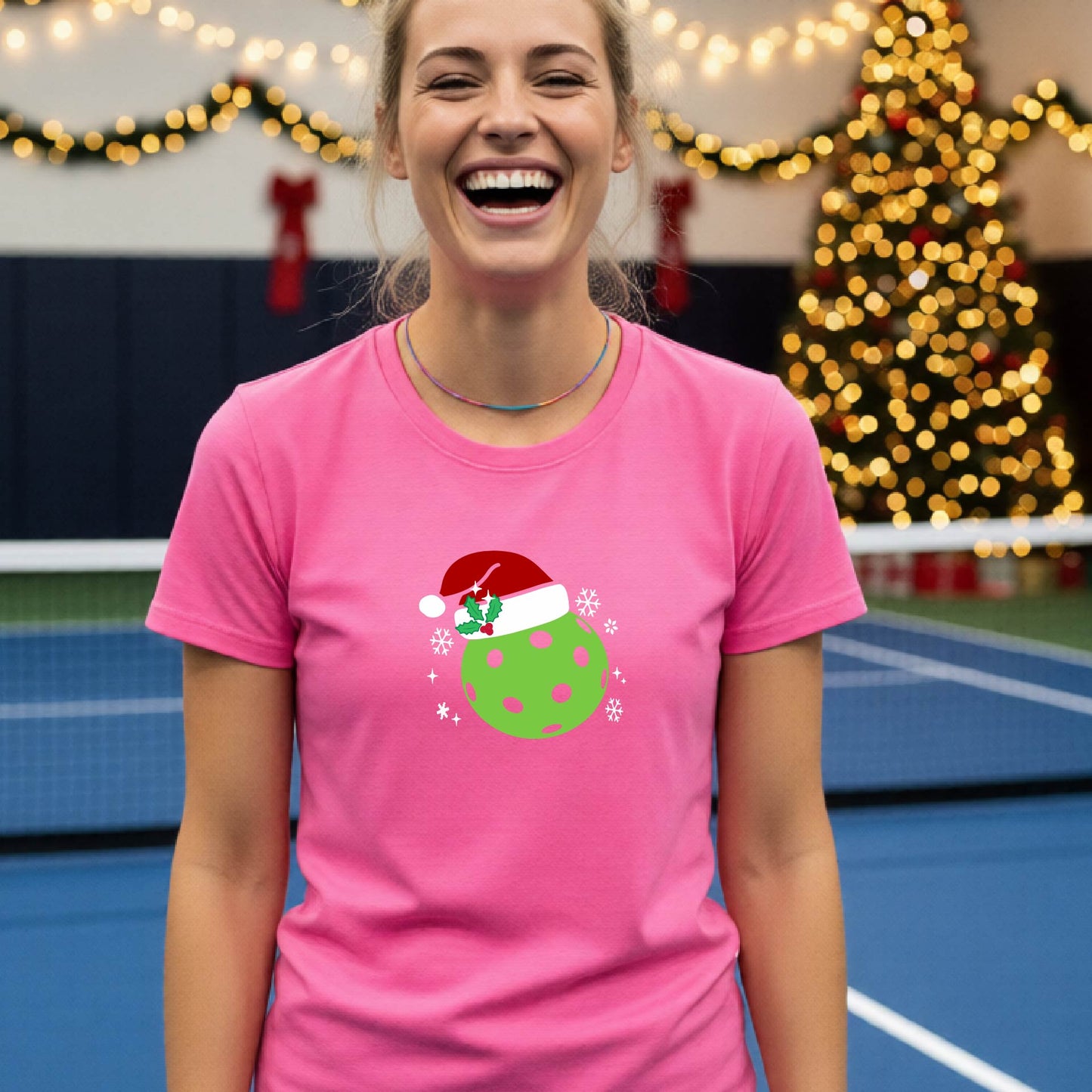 A woman wearing a bright pink graphic t-shirt with a green Christmas-themed pickleball design, standing in front of a blurred Christmas tree and holiday decorations.