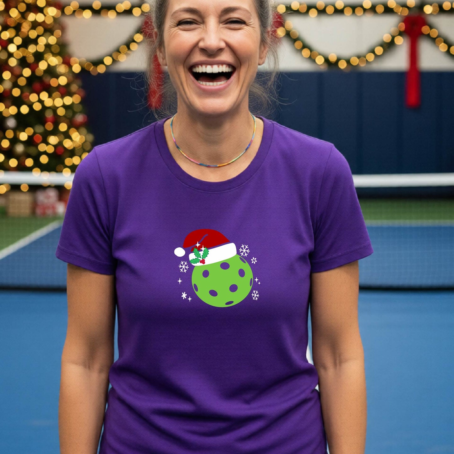 A woman wearing a purple graphic t-shirt with a green Christmas-themed pickleball design, standing in front of a blurred Christmas tree and holiday decorations.