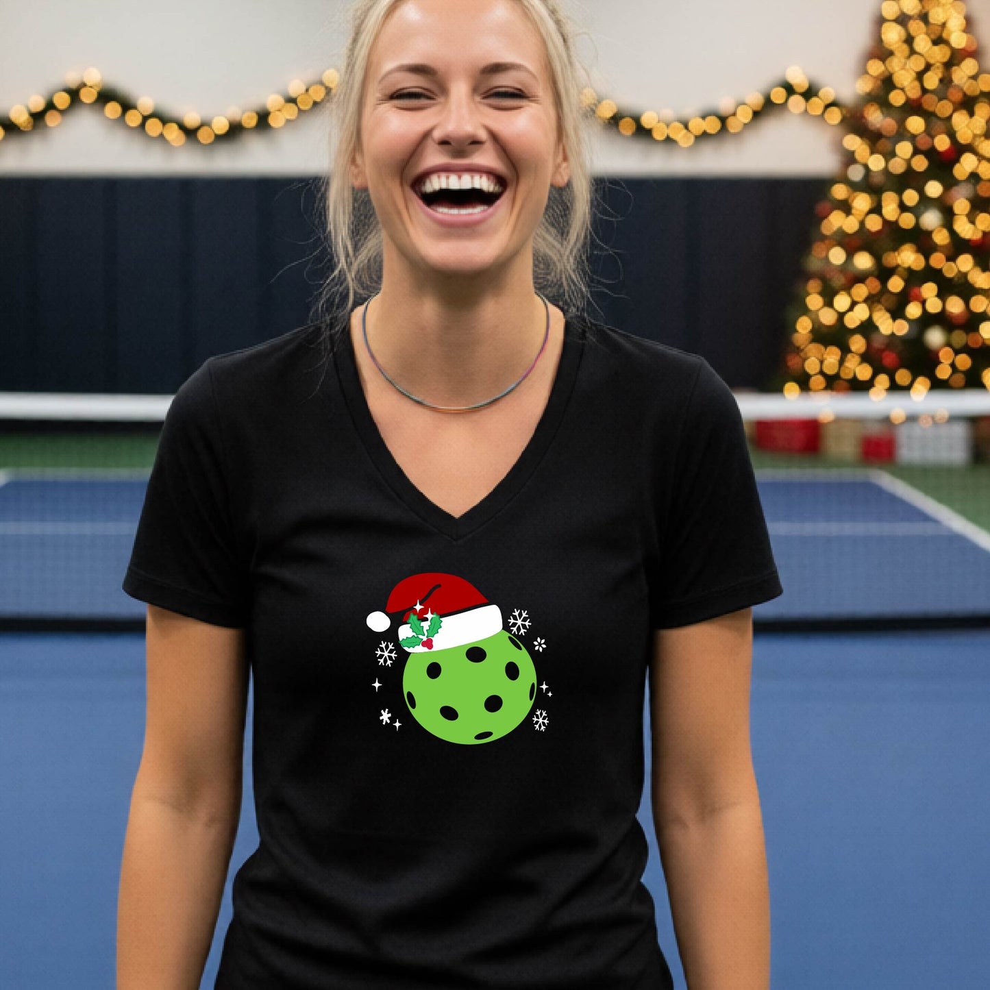 A woman smiling in a gym wearing a black v-neck t-shirt with a green Christmas pickleball santa hat graphic on the front, with holiday decorations and a Christmas tree in the background.