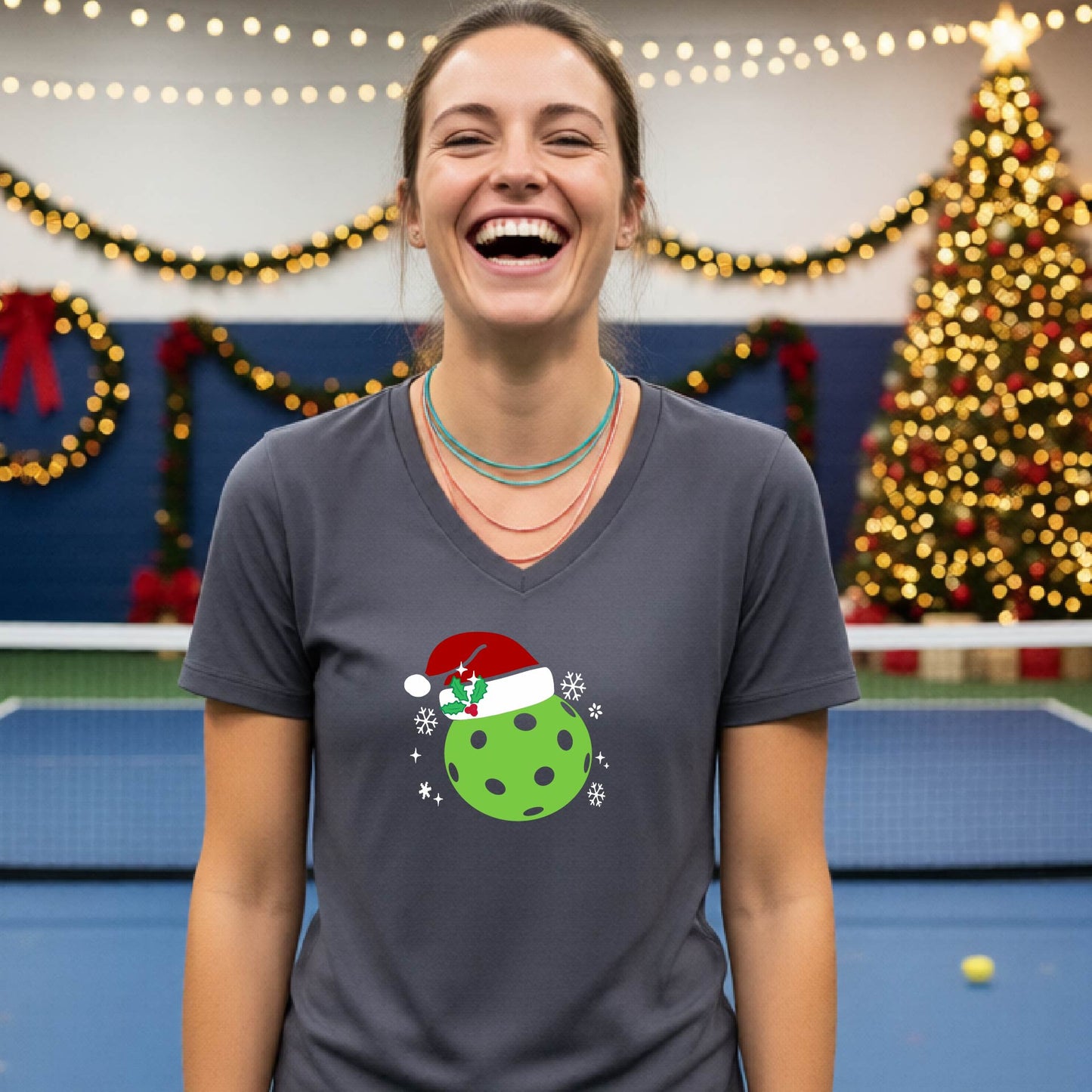 A woman smiling in a gym wearing a dark gray v-neck t-shirt with a green Christmas pickleball santa hat graphic on the front, with holiday decorations and a Christmas tree in the background.