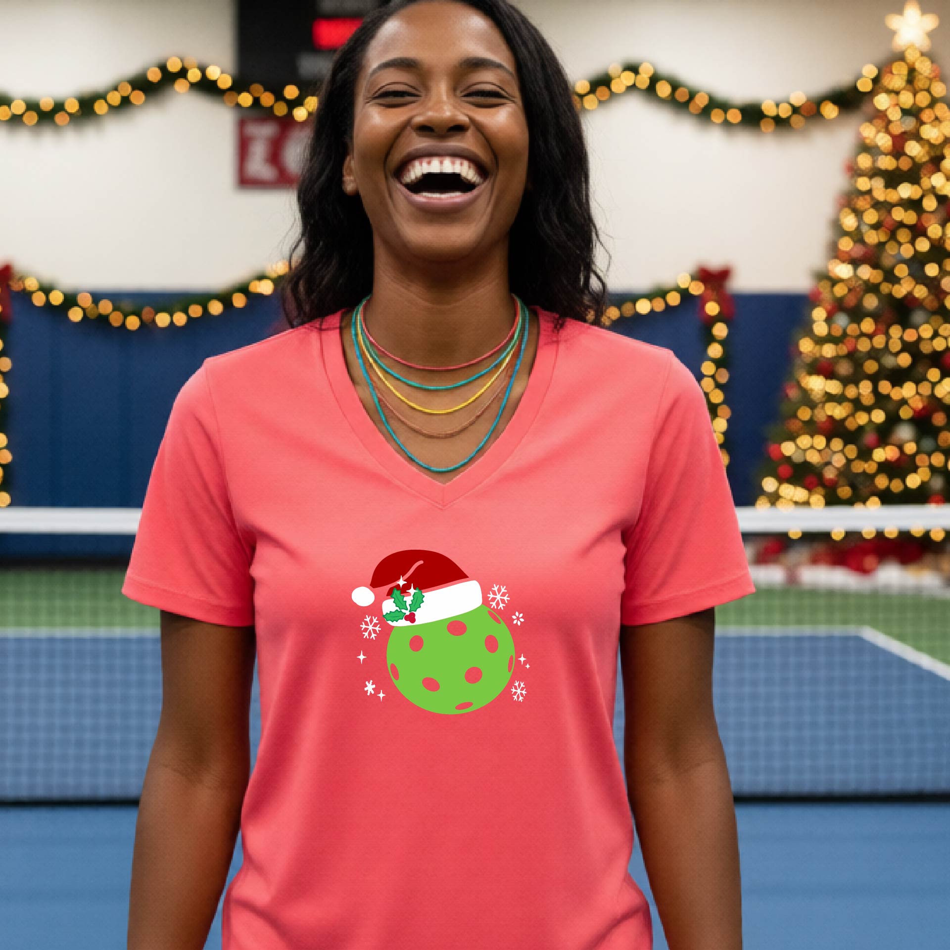 A woman smiling in a gym wearing a coral pink v-neck t-shirt with a green Christmas pickleball santa hat graphic on the front, with holiday decorations and a Christmas tree in the background.