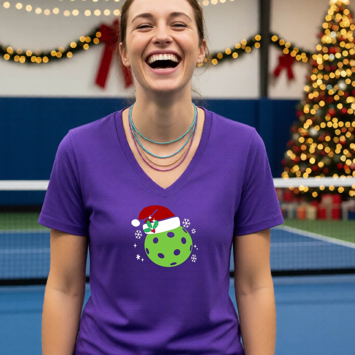 A woman smiling in a gym wearing a purple v-neck t-shirt with a green Christmas pickleball santa hat graphic on the front, with holiday decorations and a Christmas tree in the background.