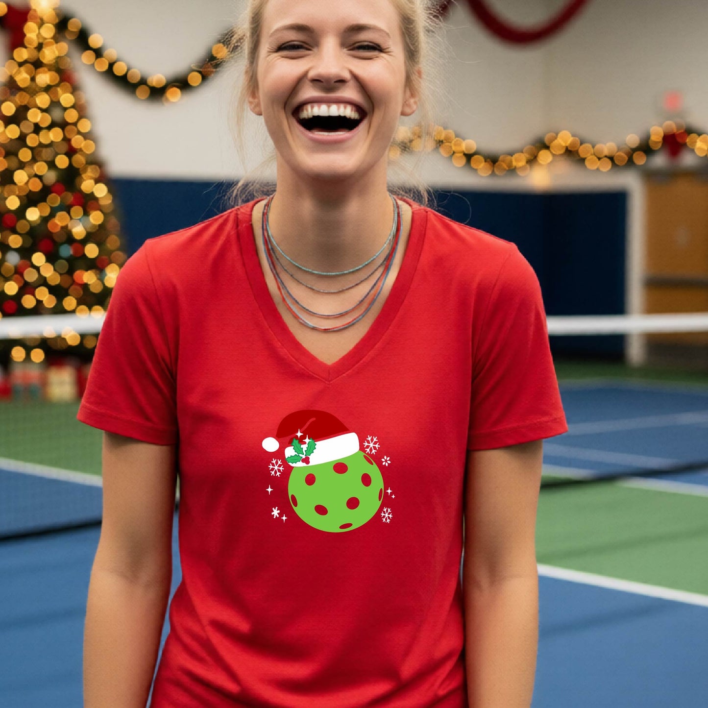 A woman smiling in a gym wearing a red v-neck t-shirt with a green Christmas pickleball santa hat graphic on the front, with holiday decorations and a Christmas tree in the background.