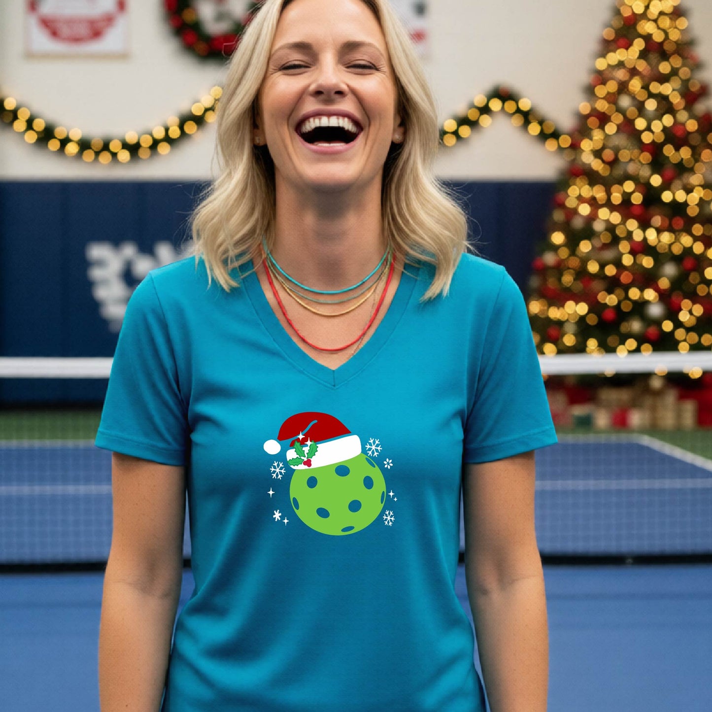 A woman smiling in a gym wearing a teal v-neck t-shirt with a green Christmas pickleball santa hat graphic on the front, with holiday decorations and a Christmas tree in the background.