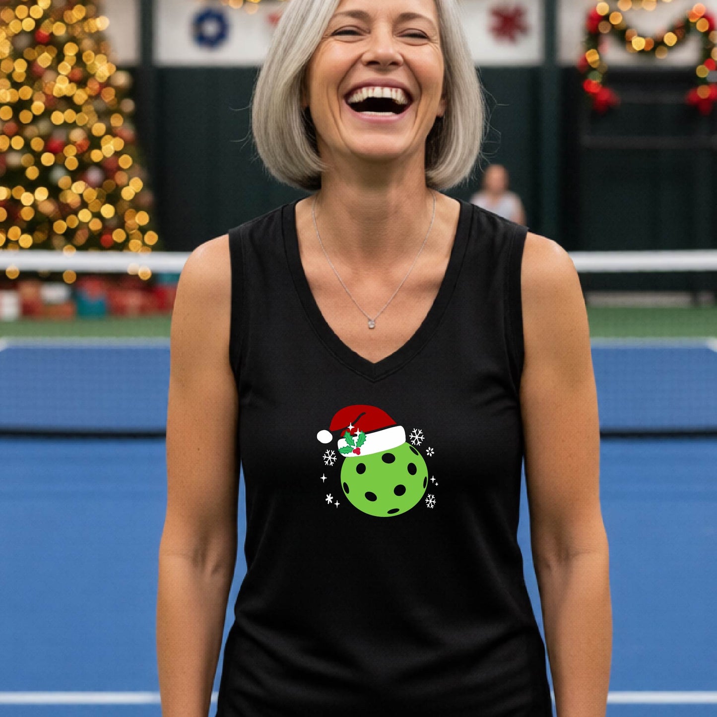 A woman laughing on an indoor pickleball court wearing a black tank top with a green Christmas pickleball ornament graphic on the chest.