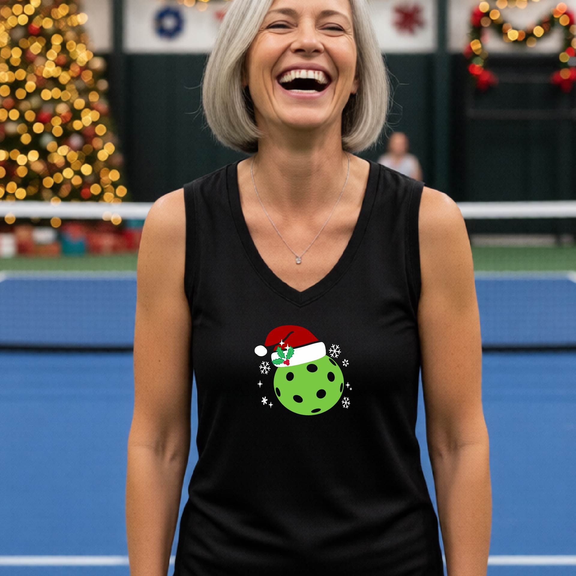A woman laughing on an indoor pickleball court wearing a black tank top with a green Christmas pickleball ornament graphic on the chest.